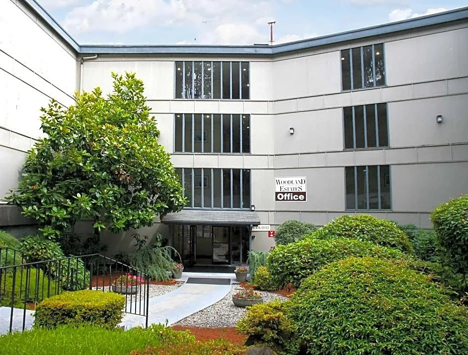 Exterior view of a modern office building with a sign that says 'Woodland Estates Office,' surrounded by lush green bushes, trees, and a landscaped pathway leading to the entrance.