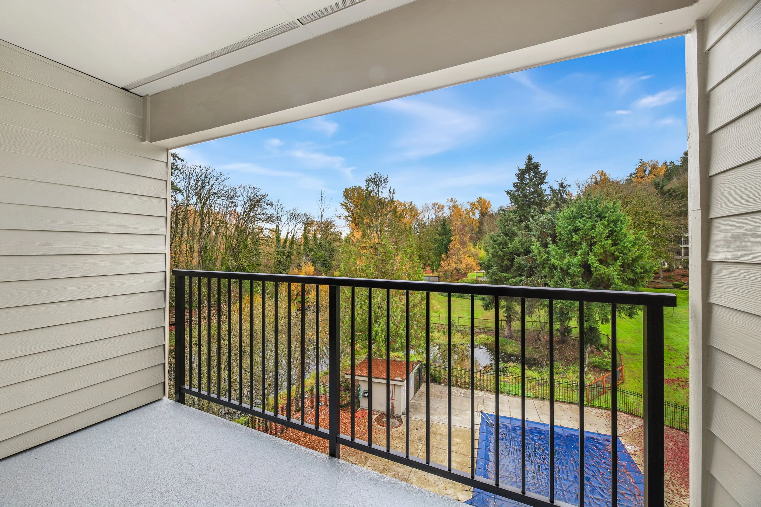 View from a balcony showing trees with autumn foliage, a small pond, a shed, a blue tarp-covered area, and green grass under a partly cloudy blue sky.