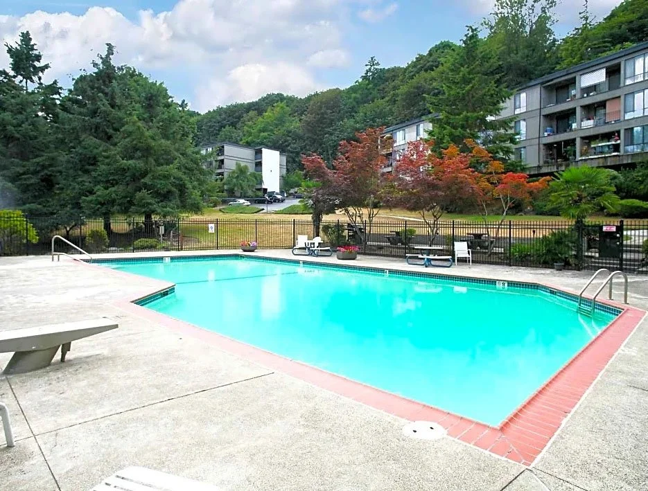 Empty outdoor swimming pool with a concrete deck, surrounded by trees and residential buildings.