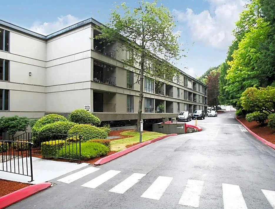 Exterior view of a multi-story residential building with parking lot and landscaped bushes, near a crosswalk on a cloudy day.