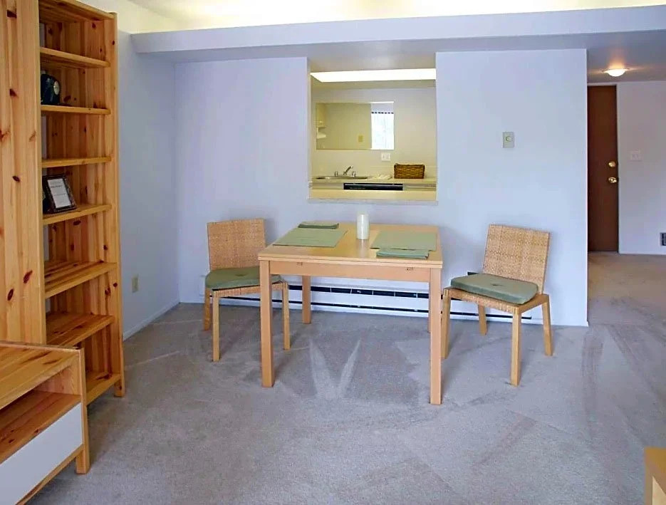 Dining area with a wooden table and two chairs with green cushions, set against a white wall with a pass-through window looking into the kitchen.
