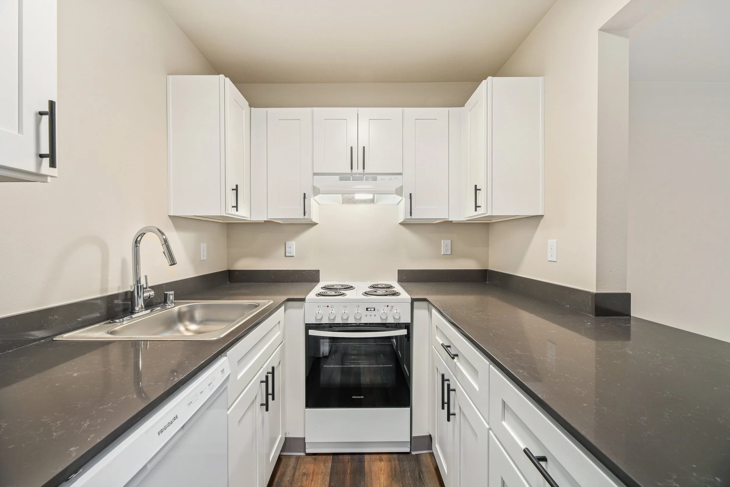 View of a small kitchen with white cabinets, a dark gray countertop, a stainless steel sink, a white electric stove, and a dishwasher, with a beige wall and wood floor.