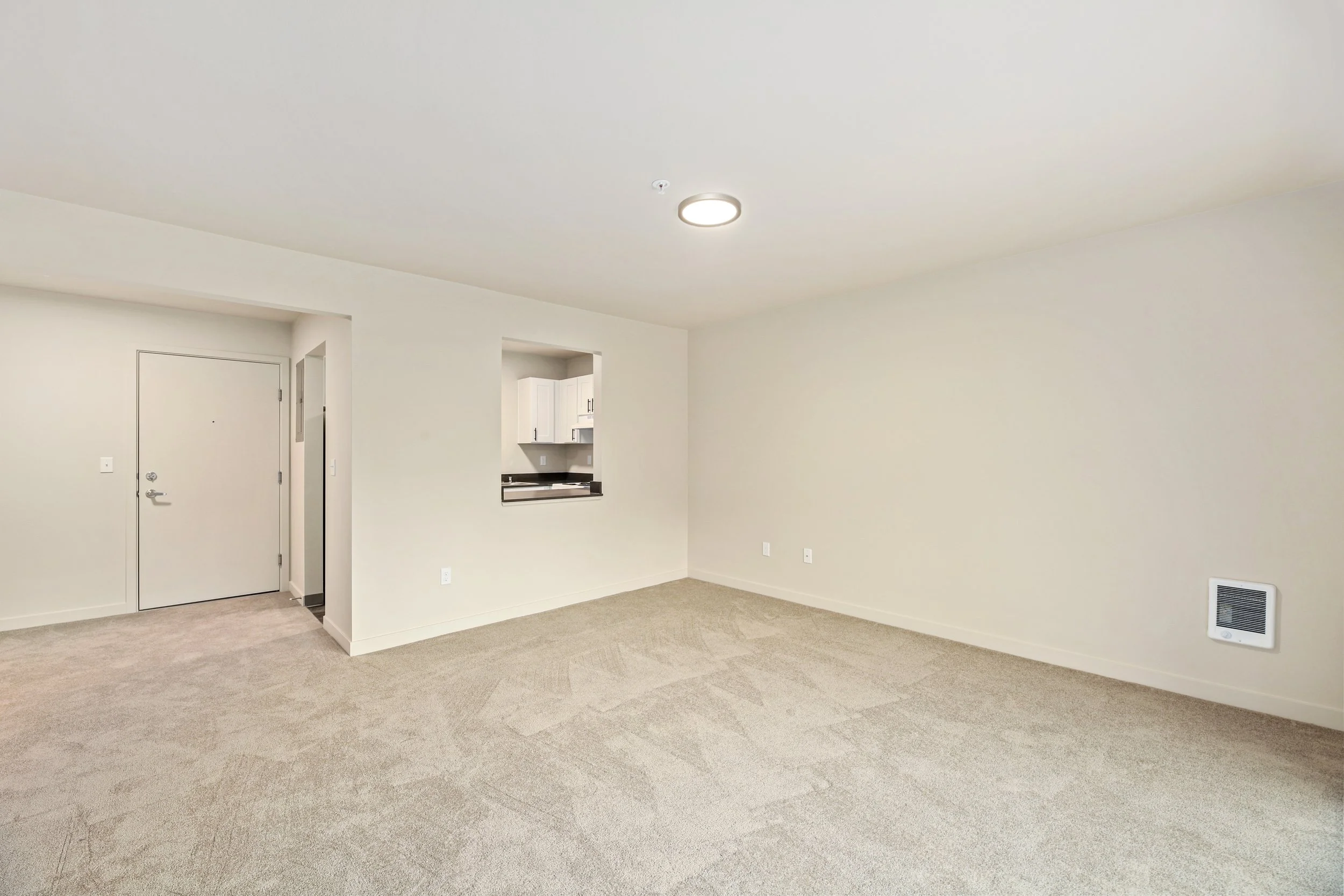 Empty living room with beige carpet, white walls, ceiling light, and a small opening to the kitchen.