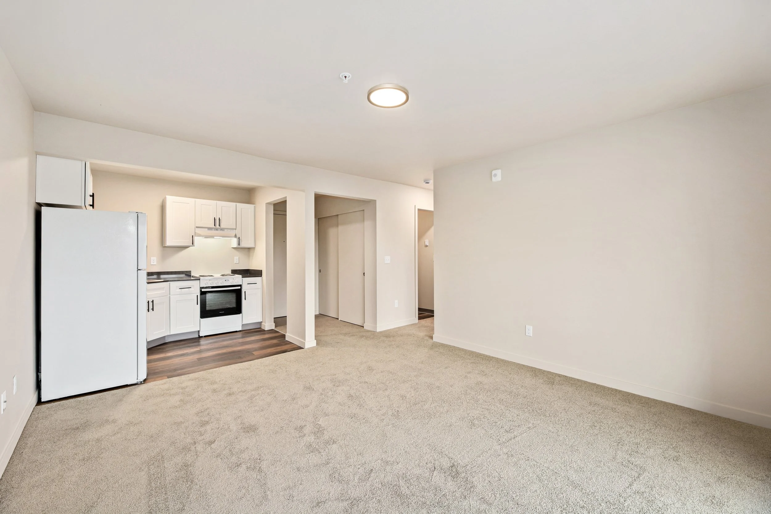 Empty living room with beige carpet and white walls, opening to a small kitchen with white cabinets, black countertops, and a white refrigerator, in an apartment or condo.
