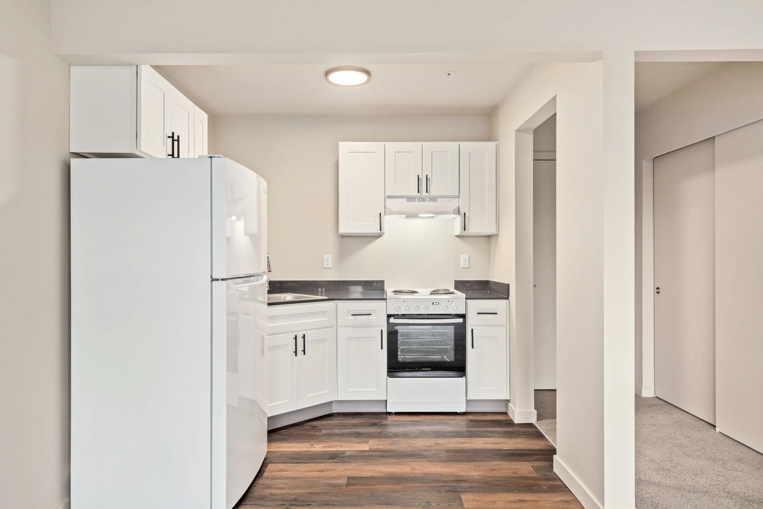 A small kitchen with white cabinets, black countertops, a white refrigerator, a white stove, and a stainless steel sink. The floor is wood, and there is a circular ceiling light.
