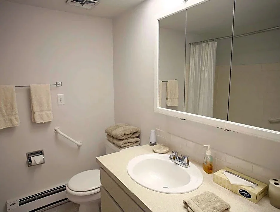 A bathroom with a white sink, countertop, large mirror, and a window. There are towels on a towel rack and a toilet with a stack of towels beside it.
