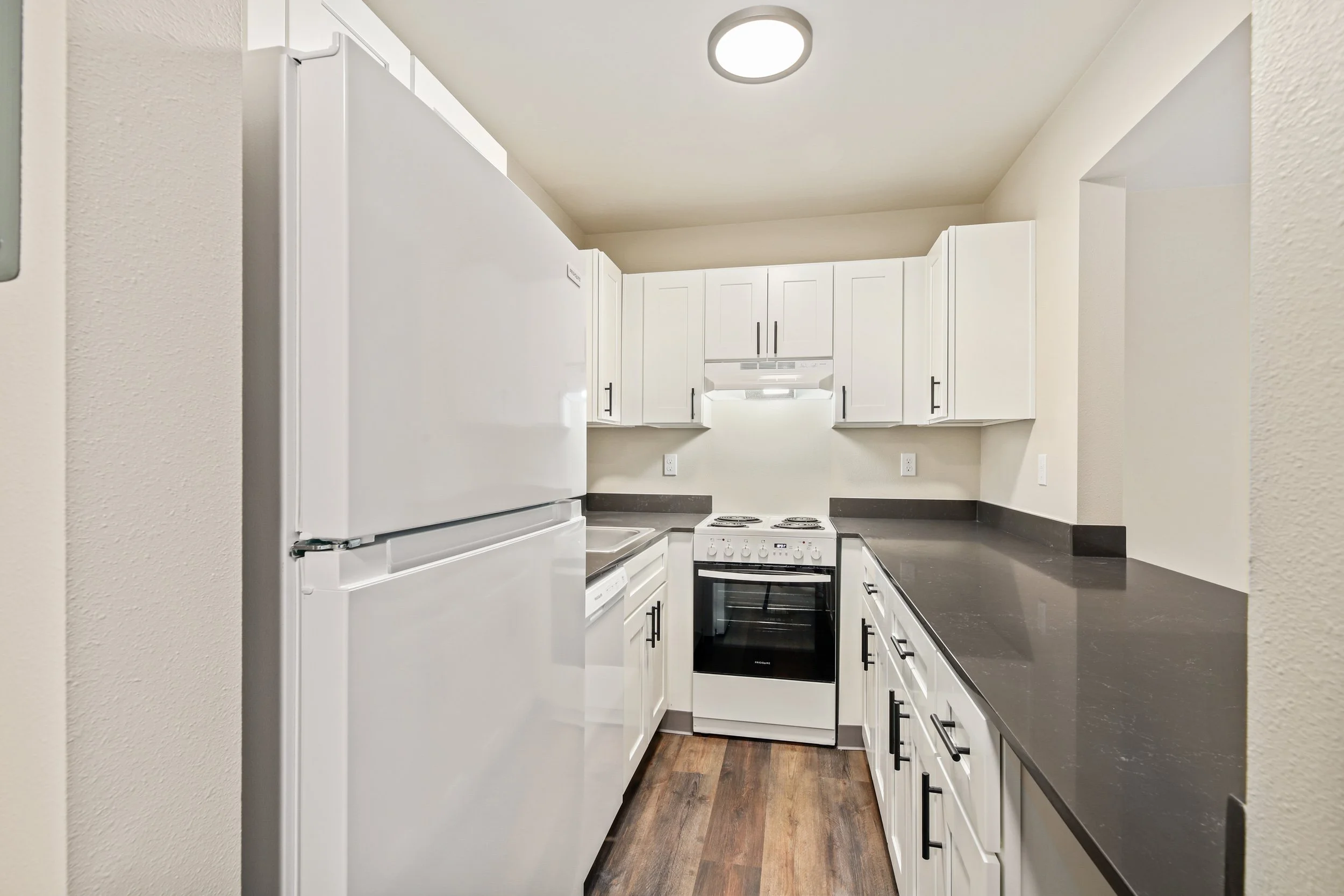 Small kitchen with white cabinets, black countertops, white stove, and refrigerator, and a wood floor. No people or other objects visible.