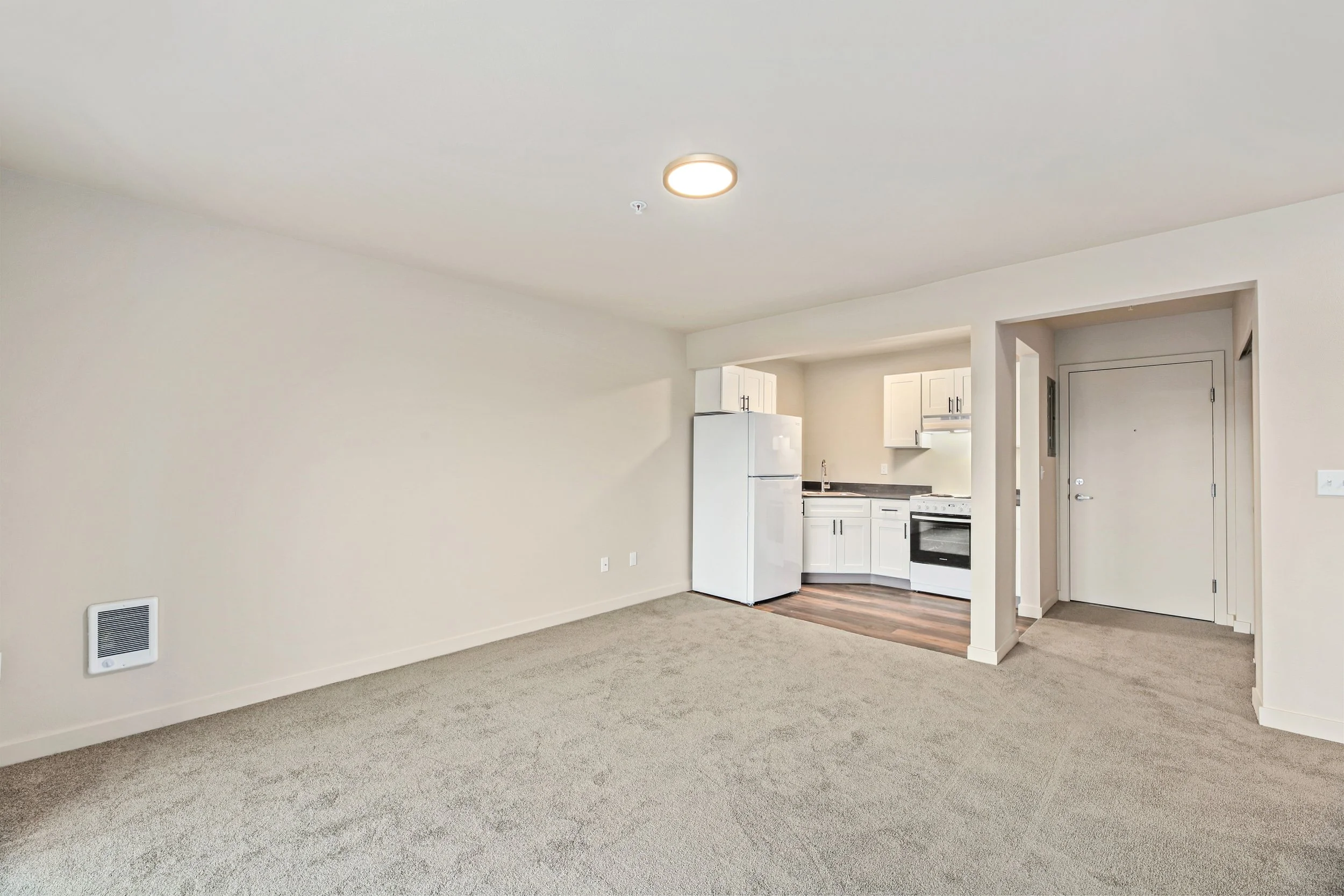 Empty living room with beige carpet, white walls, and an open kitchen with white cabinets, refrigerator, stove, and dark countertops.