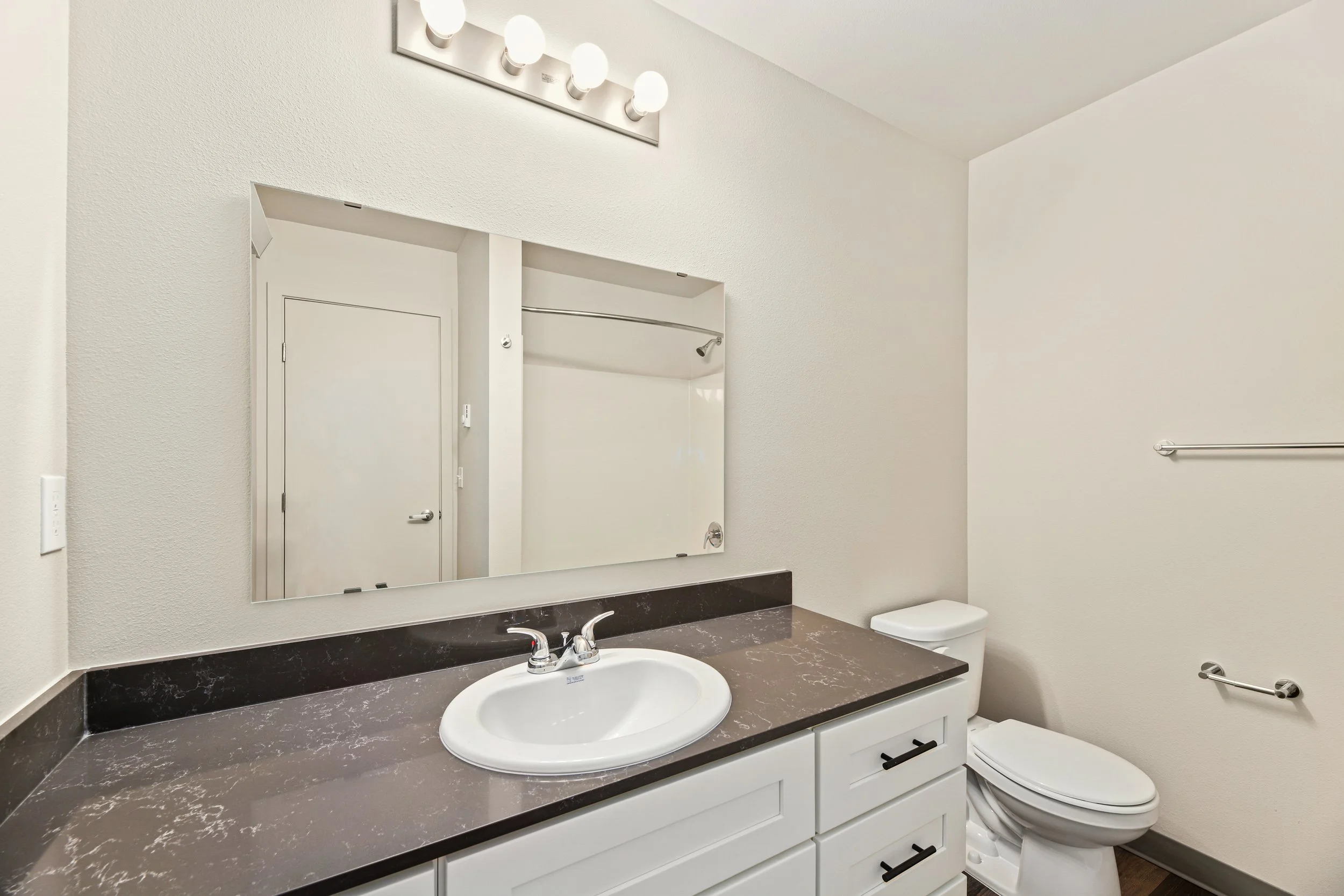 Bathroom with a dark marble countertop, a white sink with silver faucet, a large mirror, white vanity cabinets, a toilet, and empty towel racks on the wall.