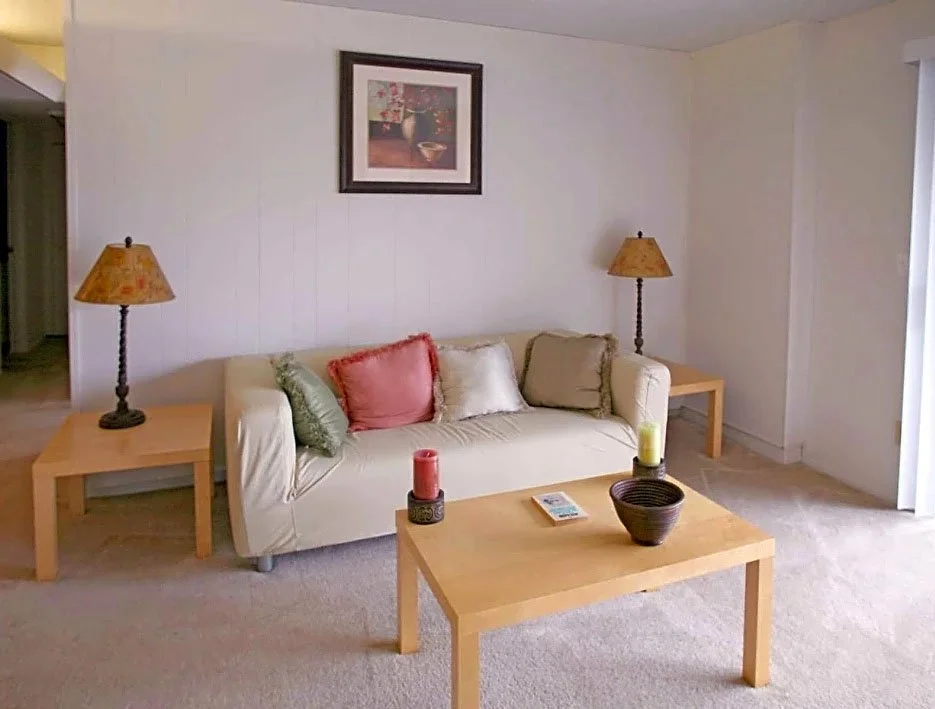 Living room with beige couch, three colorful pillows, two wooden side tables with lamps, a framed picture on the wall, and a wooden coffee table with candles, a bowl, and a magazine.