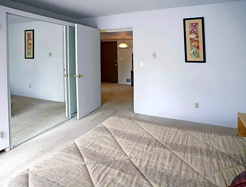 Bedroom with beige bedspread, white walls, framed artwork, mirrored closet doors, and open door leading to a hallway with a light fixture.