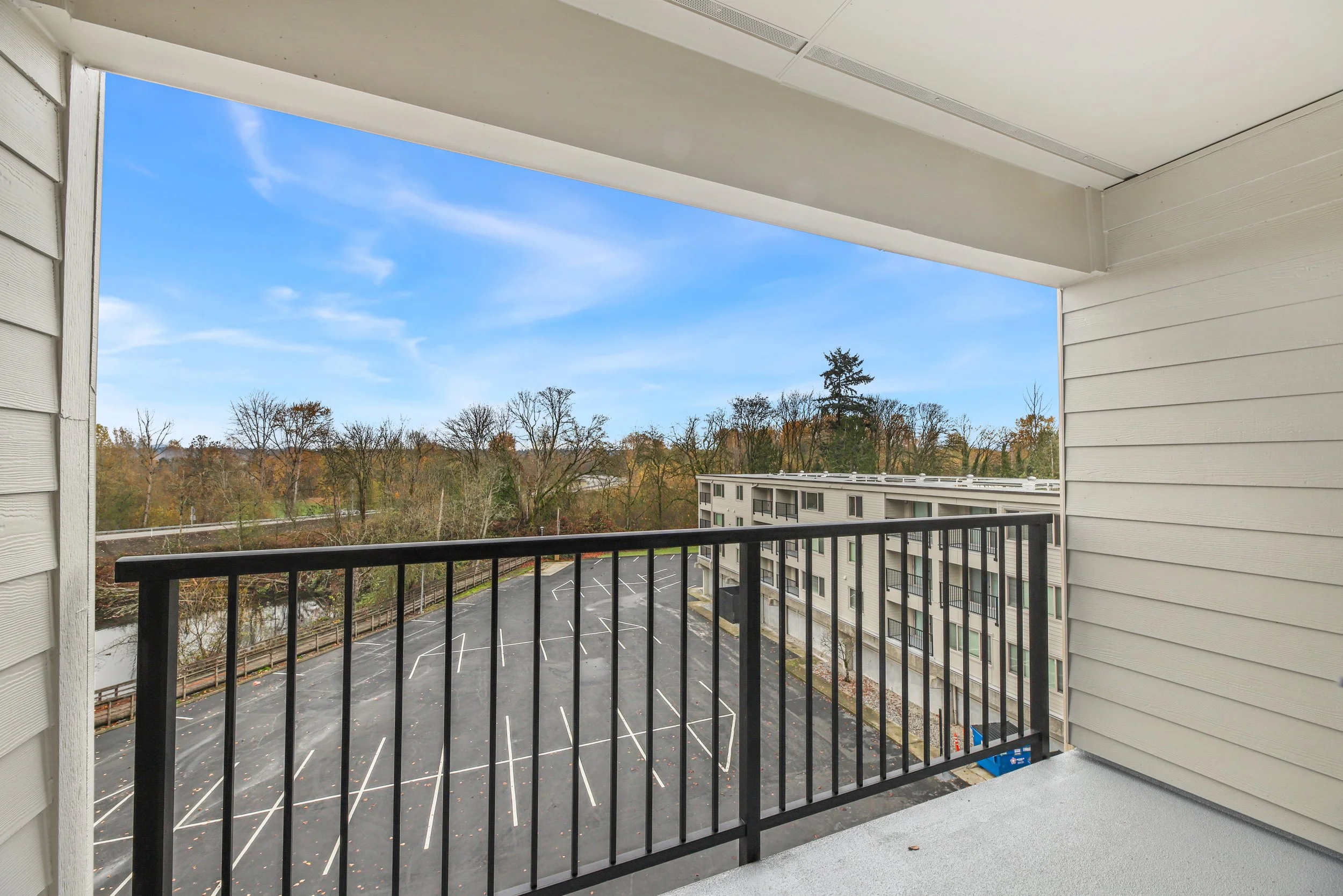 Balcony overlooking a parking lot, apartment building, and trees with autumn foliage under a blue sky.