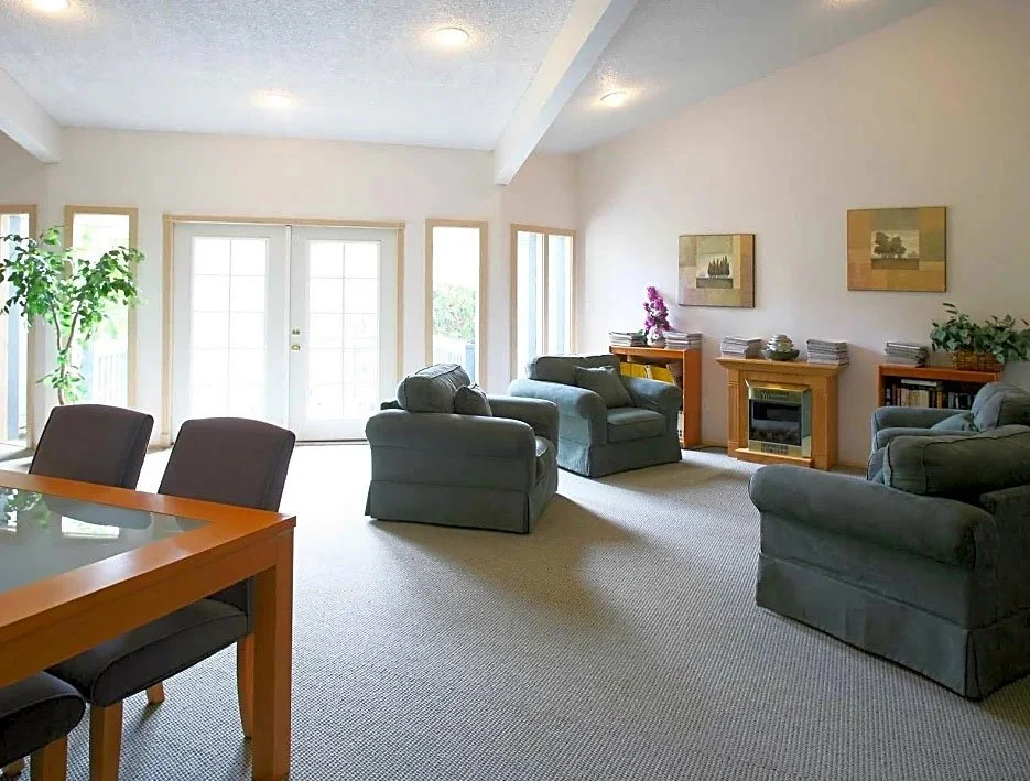 A spacious living room with beige walls, large windows, a sliding glass door, four dark green armchairs, a wooden fireplace mantel, a plant on the left, and two framed landscape paintings on the wall.