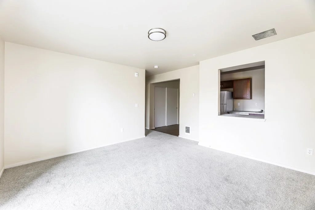 Empty living room with carpeted floor, white walls, ceiling light, view into a kitchen with wooden cabinets and refrigerator through a wall opening, doorway leading to other rooms.