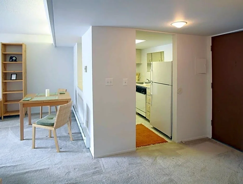 Interior view of a small apartment showing a dining area with two chairs and a table, and a kitchen with a refrigerator and stove, with plain white walls and carpeted floors.