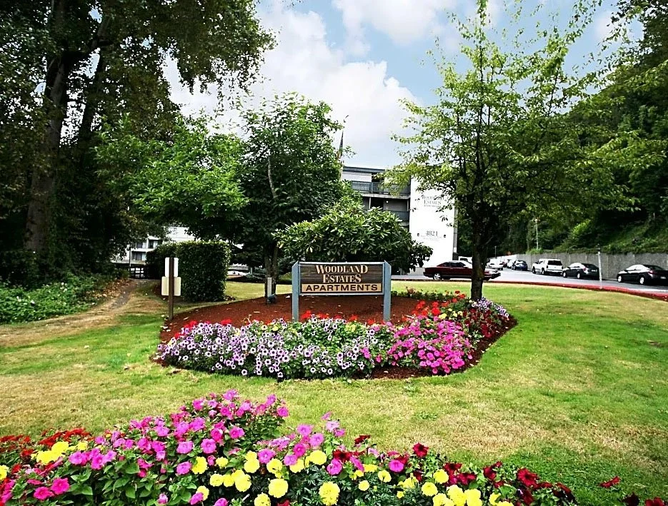 Landscaped area with colorful flowers, green grass, trees, a wooden sign reading 'Woodland Estates Apartments,' and a parking lot with cars in the background.