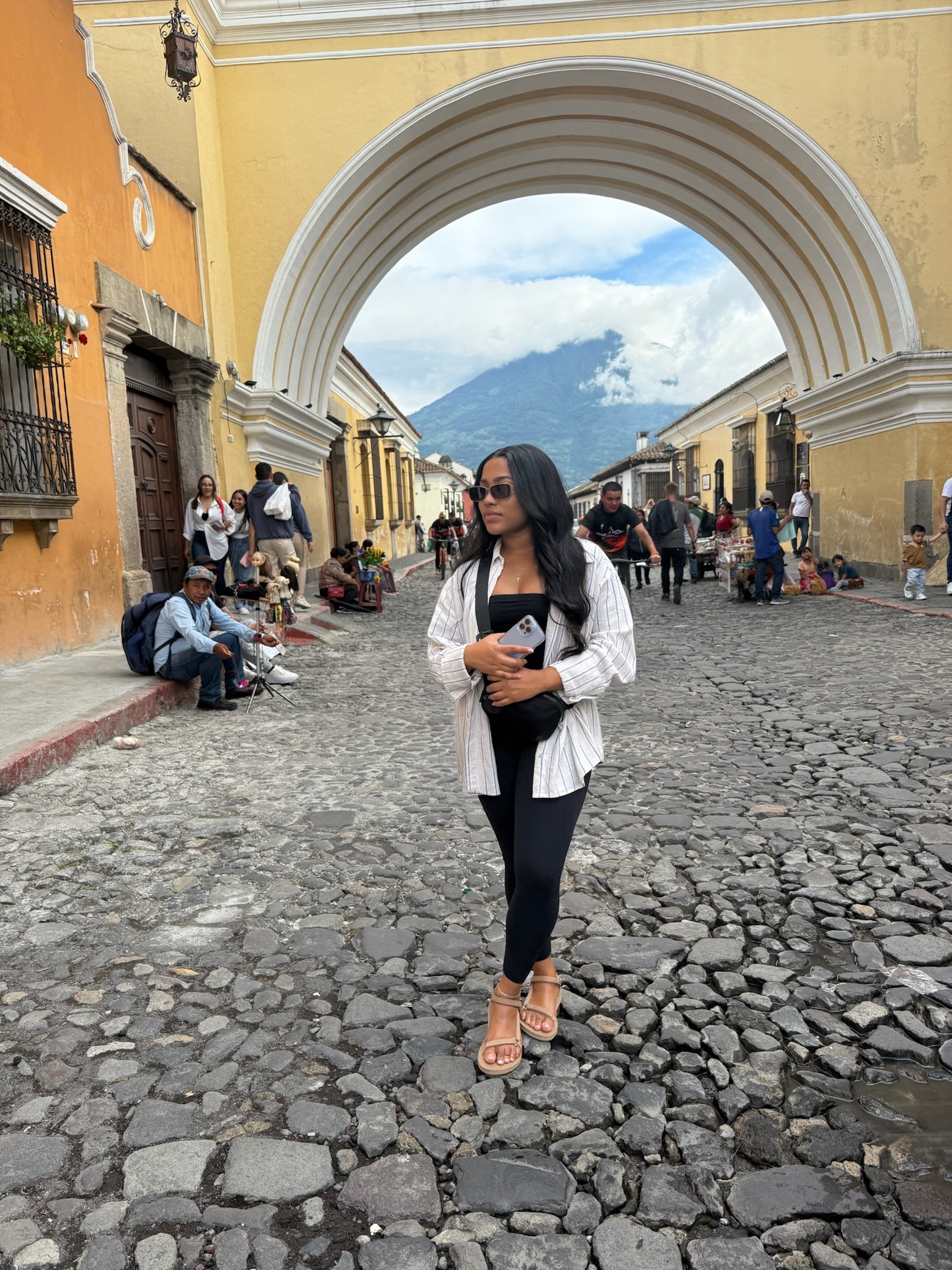 A woman with long black hair and sunglasses standing on a cobblestone street in front of a white archway with a mountain in the background. She is holding a phone and wearing a striped shirt, black leggings, and beige high-heeled sandals. People are 