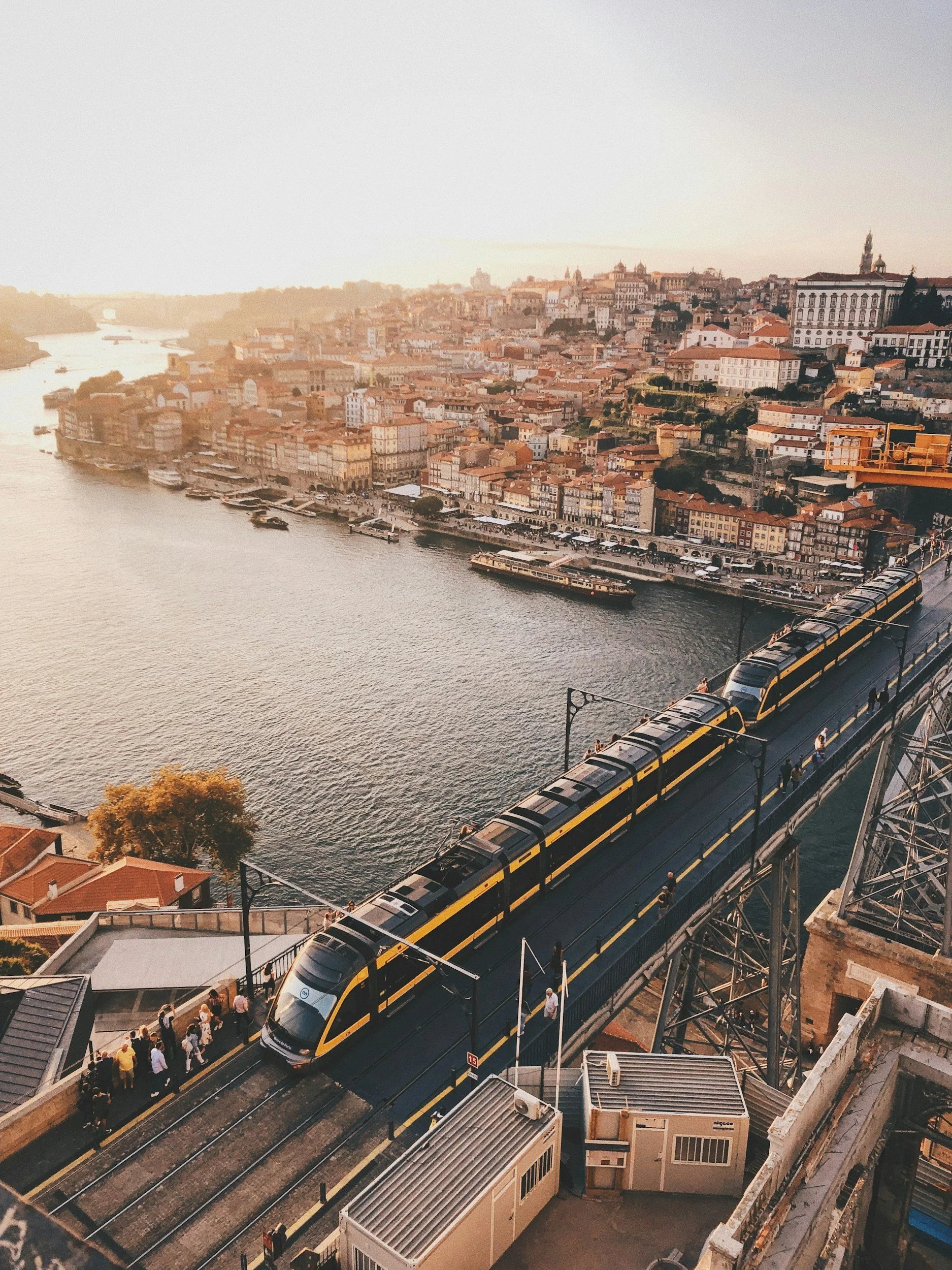 A yellow and black tram moving across a bridge over a river, with a cityscape of tightly packed buildings on a hillside in the background during sunset.