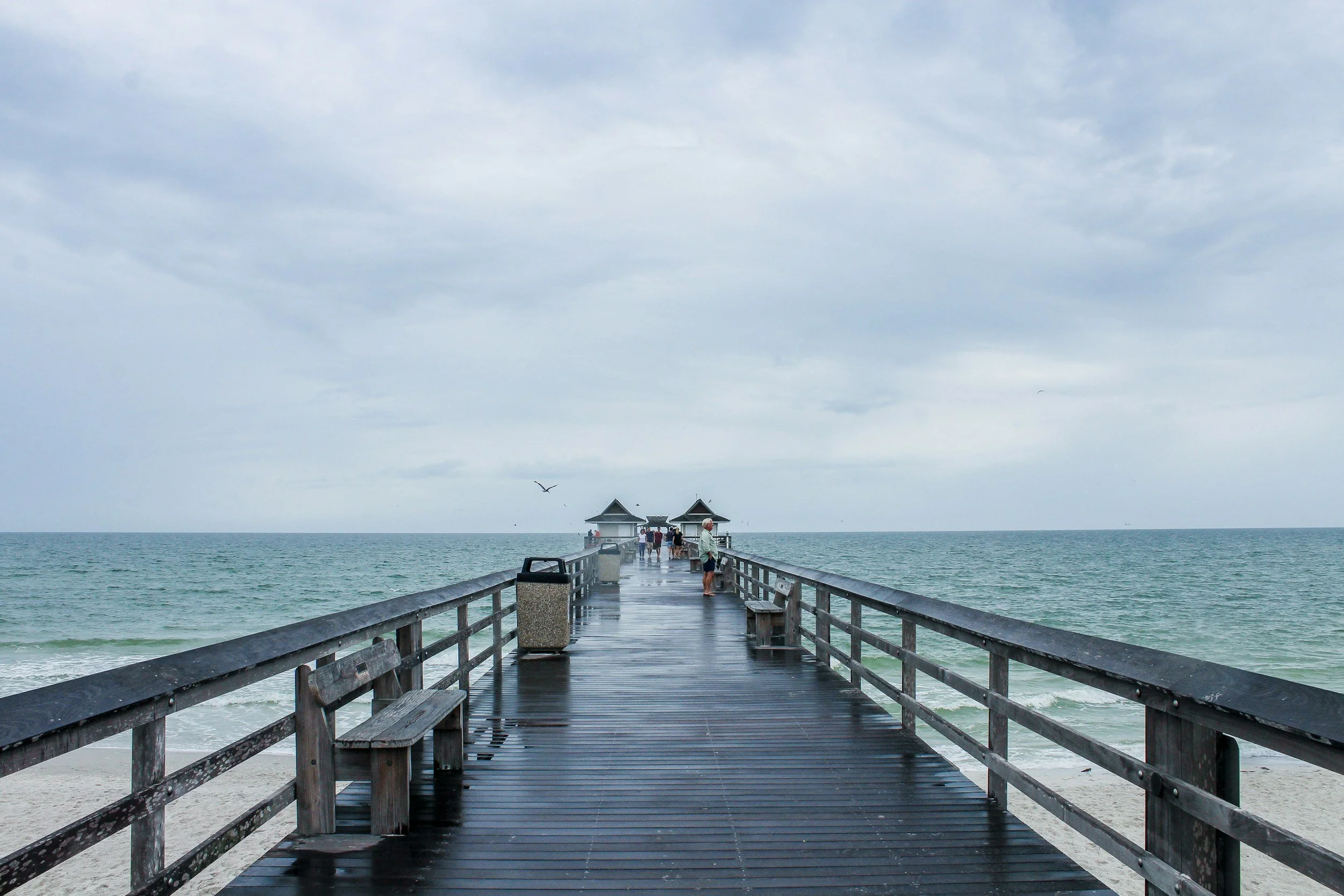Wooden pier extending into the ocean with a few people walking and sitting, cloudy sky overhead, seagulls flying around, and the water on either side of the pier.
