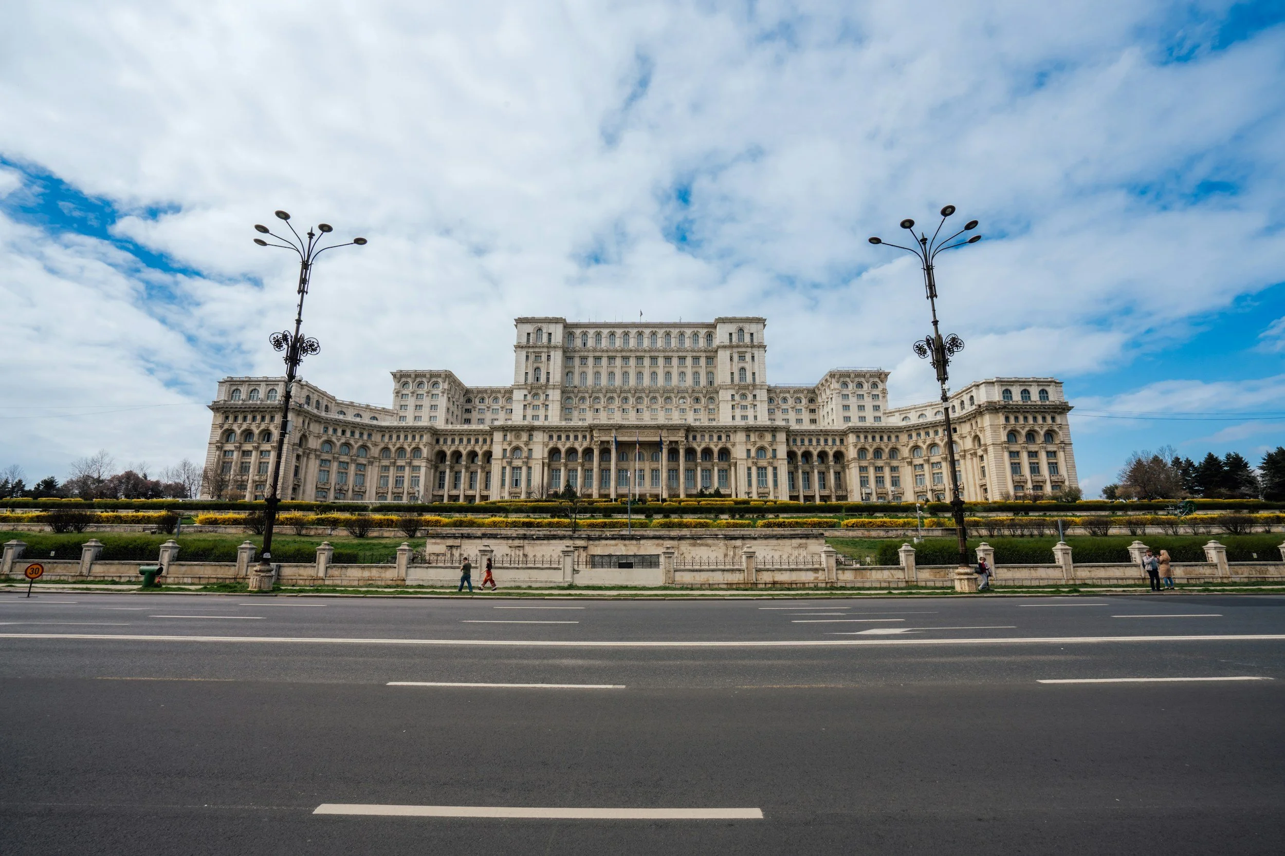 The Palace of the Parliament, large neoclassical building with multiple stories, ornate details, situated behind a wide street with street lamps, and a partly cloudy sky in the background.