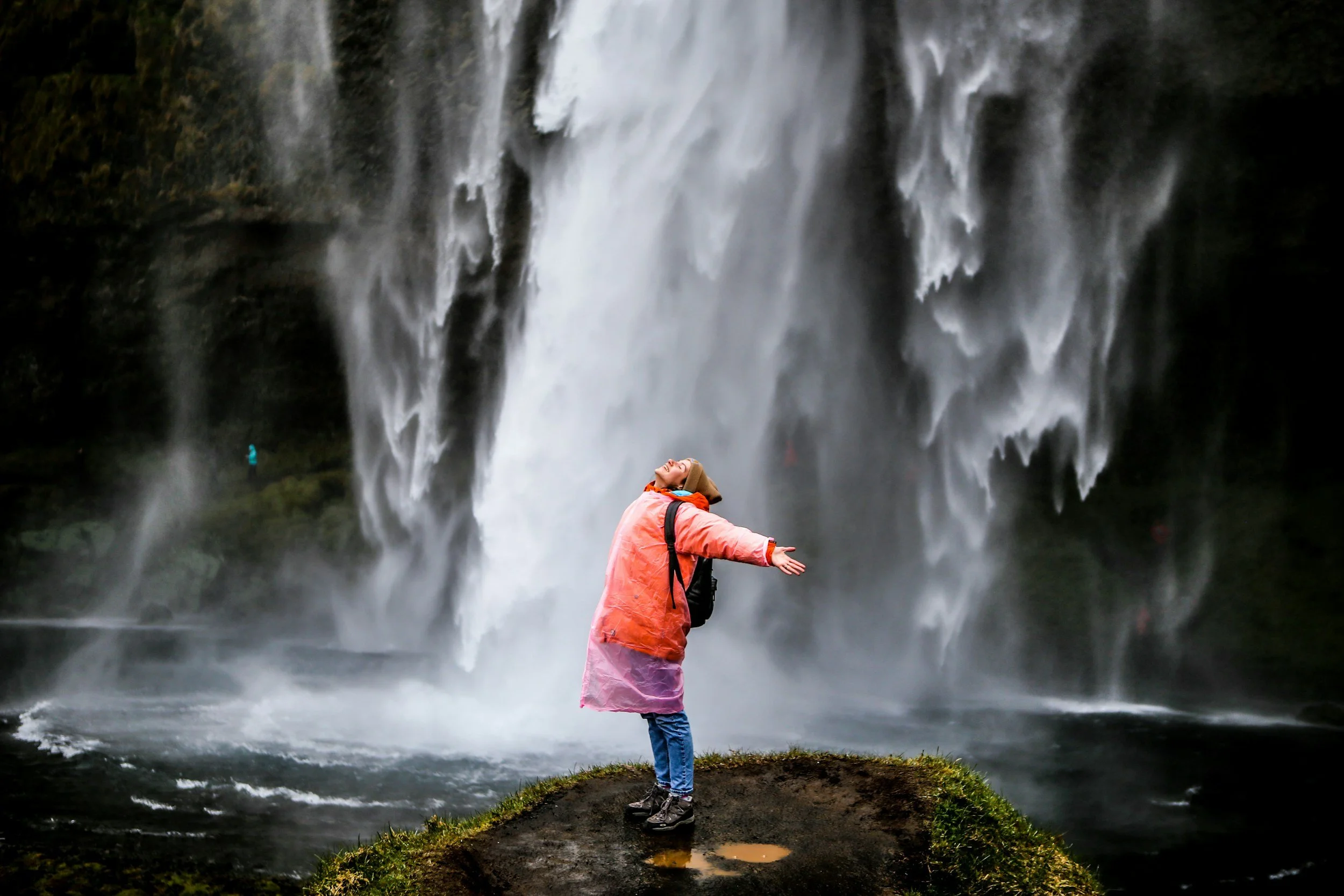 Person in pink and orange raincoat standing on a small patch of land in front of a large waterfall, with arms outstretched and head tilted back.