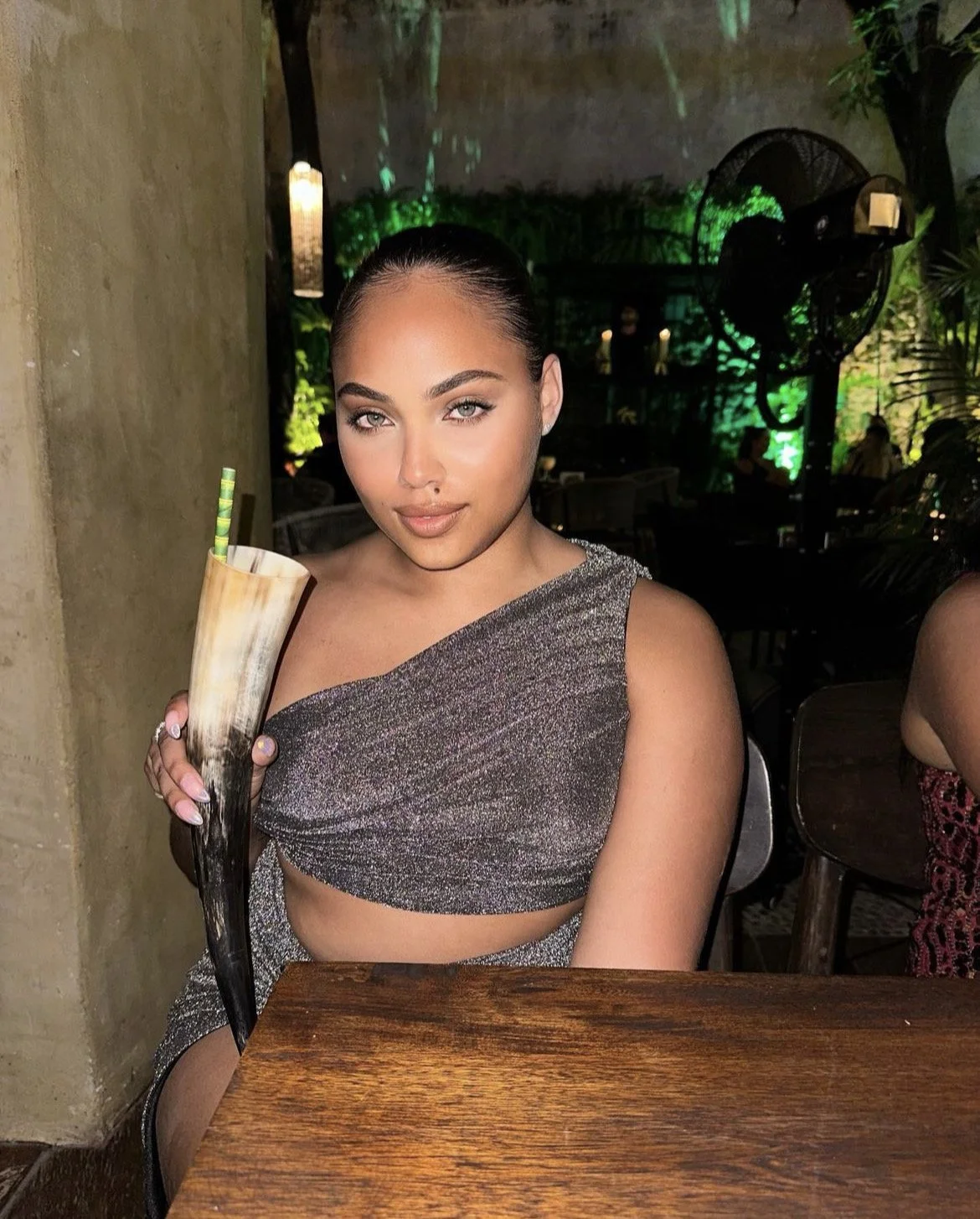 A woman with dark hair pulled back, wearing a sparkly gray crop top, sitting at a wooden table in a dimly lit restaurant or bar, holding a large curved horn-shaped drink with a striped straw.