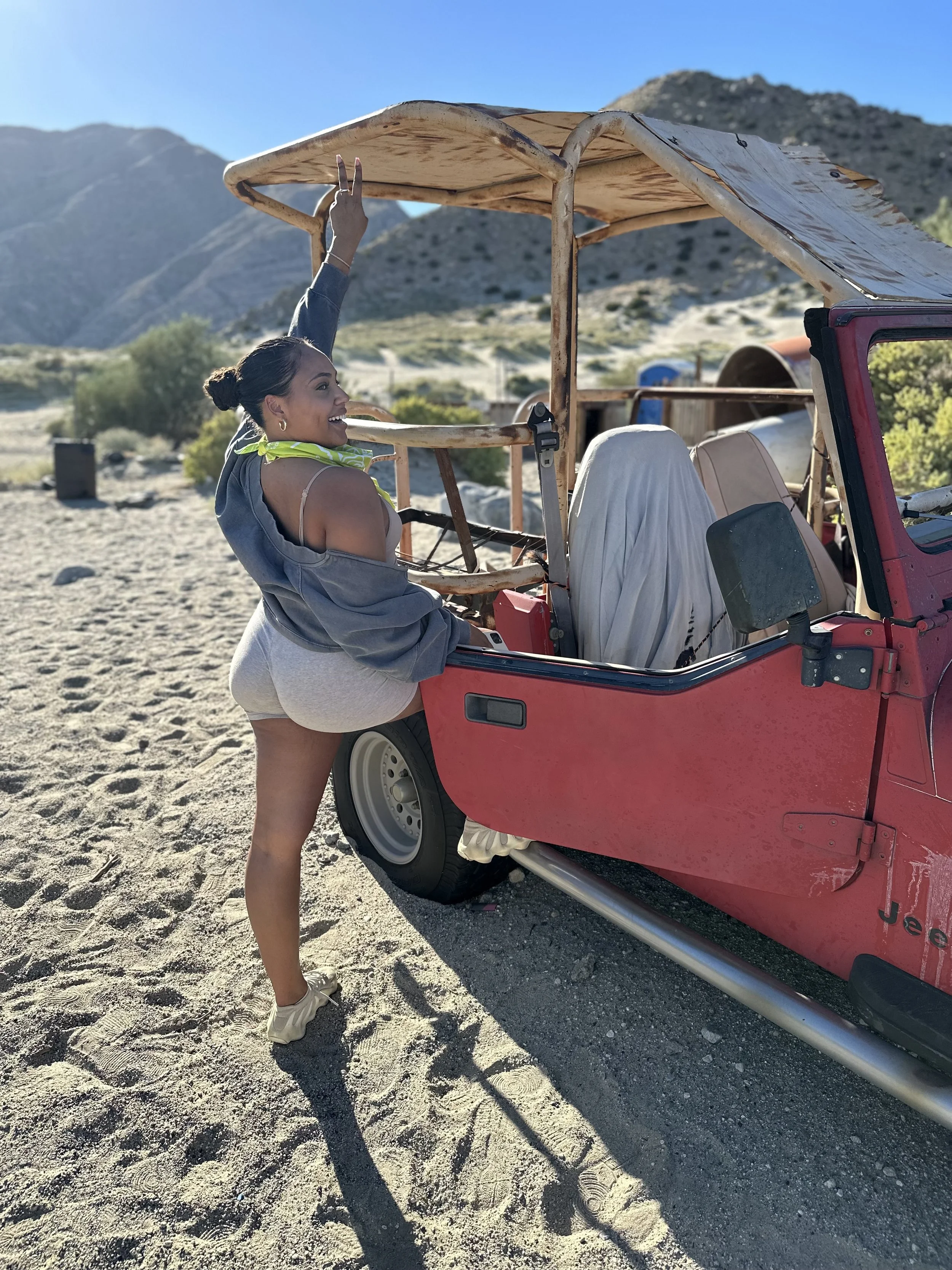 Smiling woman standing on sandy area next to a vintage off-road vehicle, holding onto the roof with one hand, in a mountainous landscape on a sunny day.