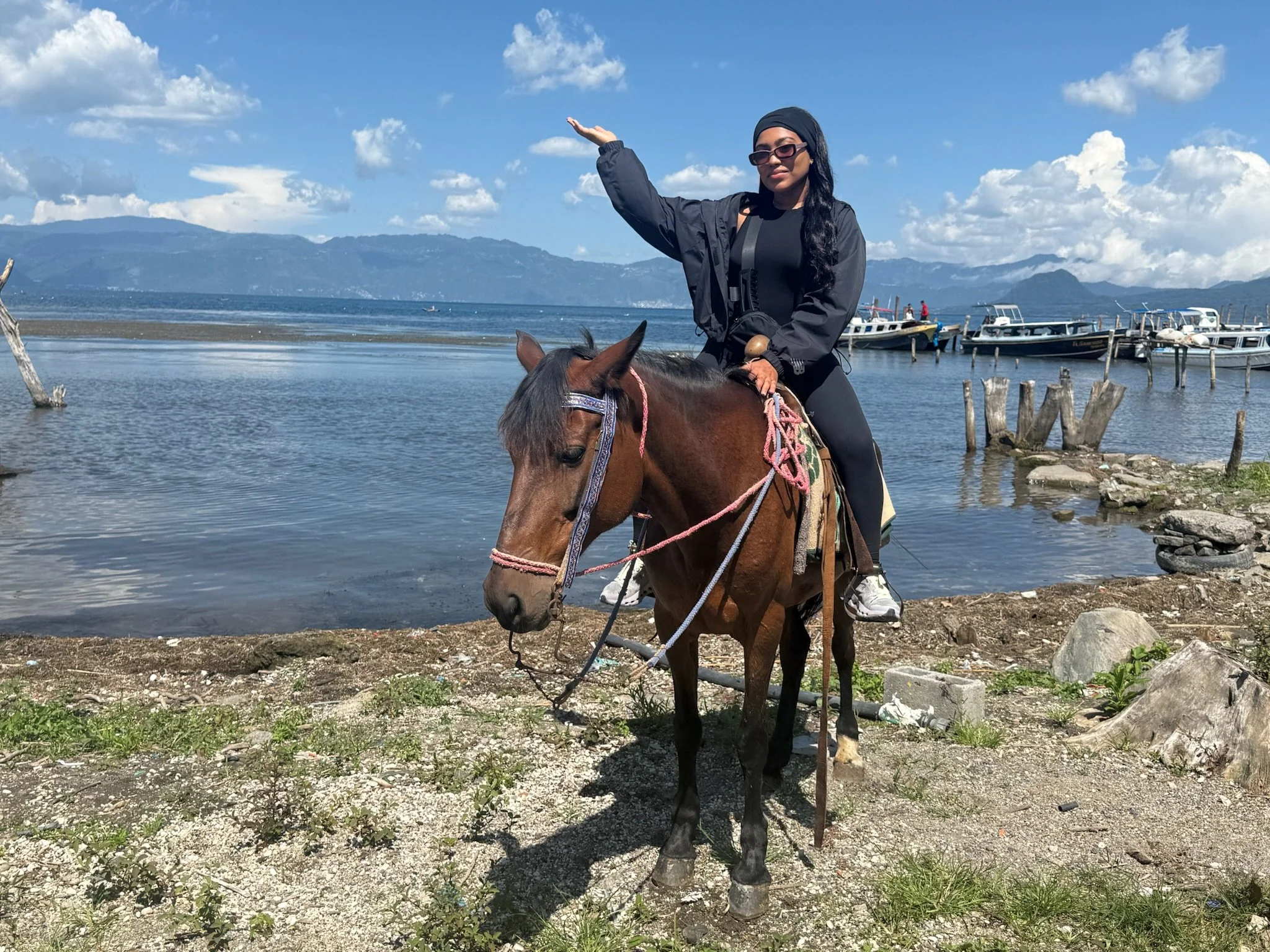 A woman riding a brown horse along a lakeside with boats docked in the background and mountains under a partly cloudy sky.
