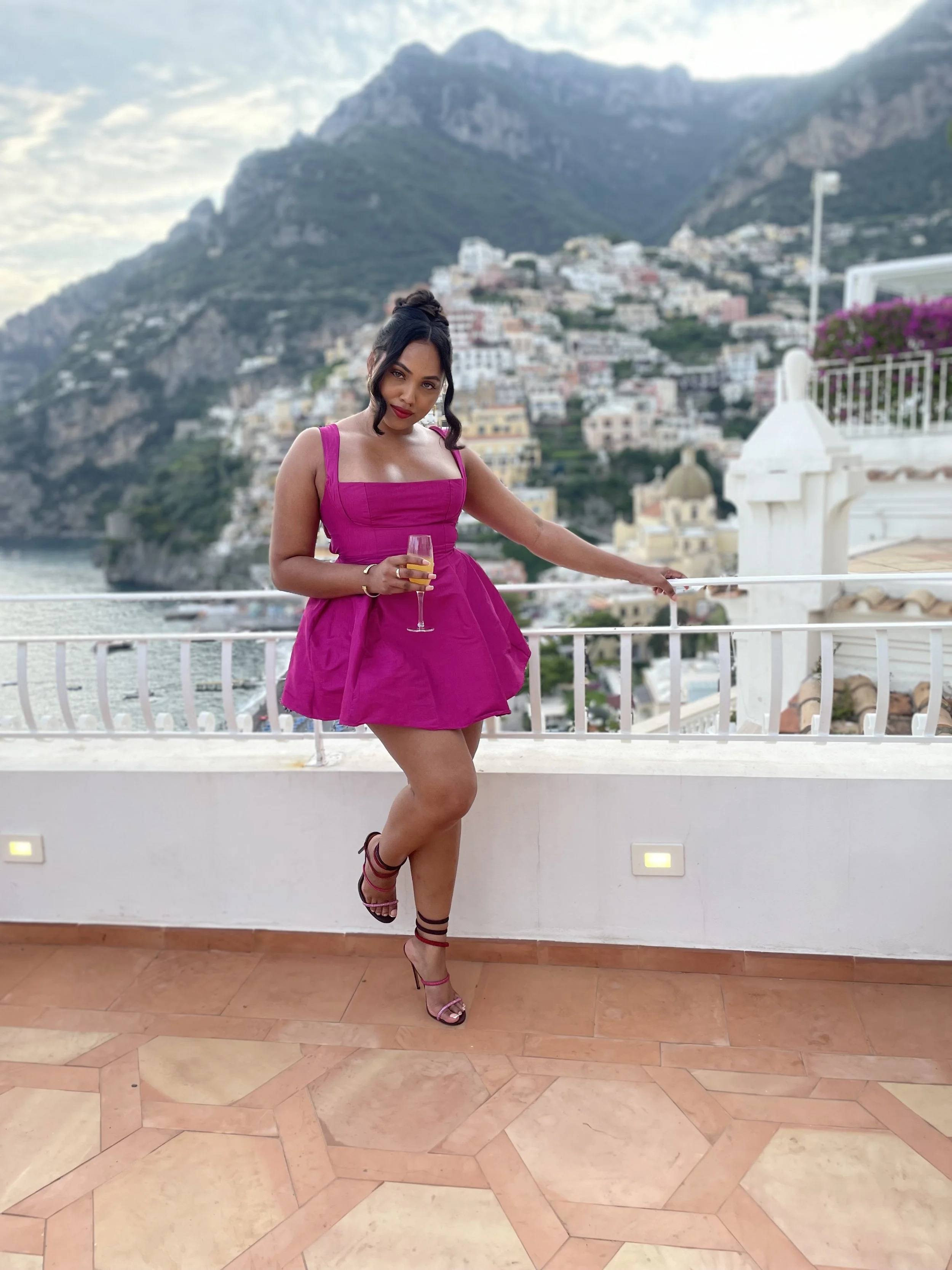 A woman in a bright pink dress holding a glass of champagne, standing on a balcony with a scenic coastal town and mountains in the background.