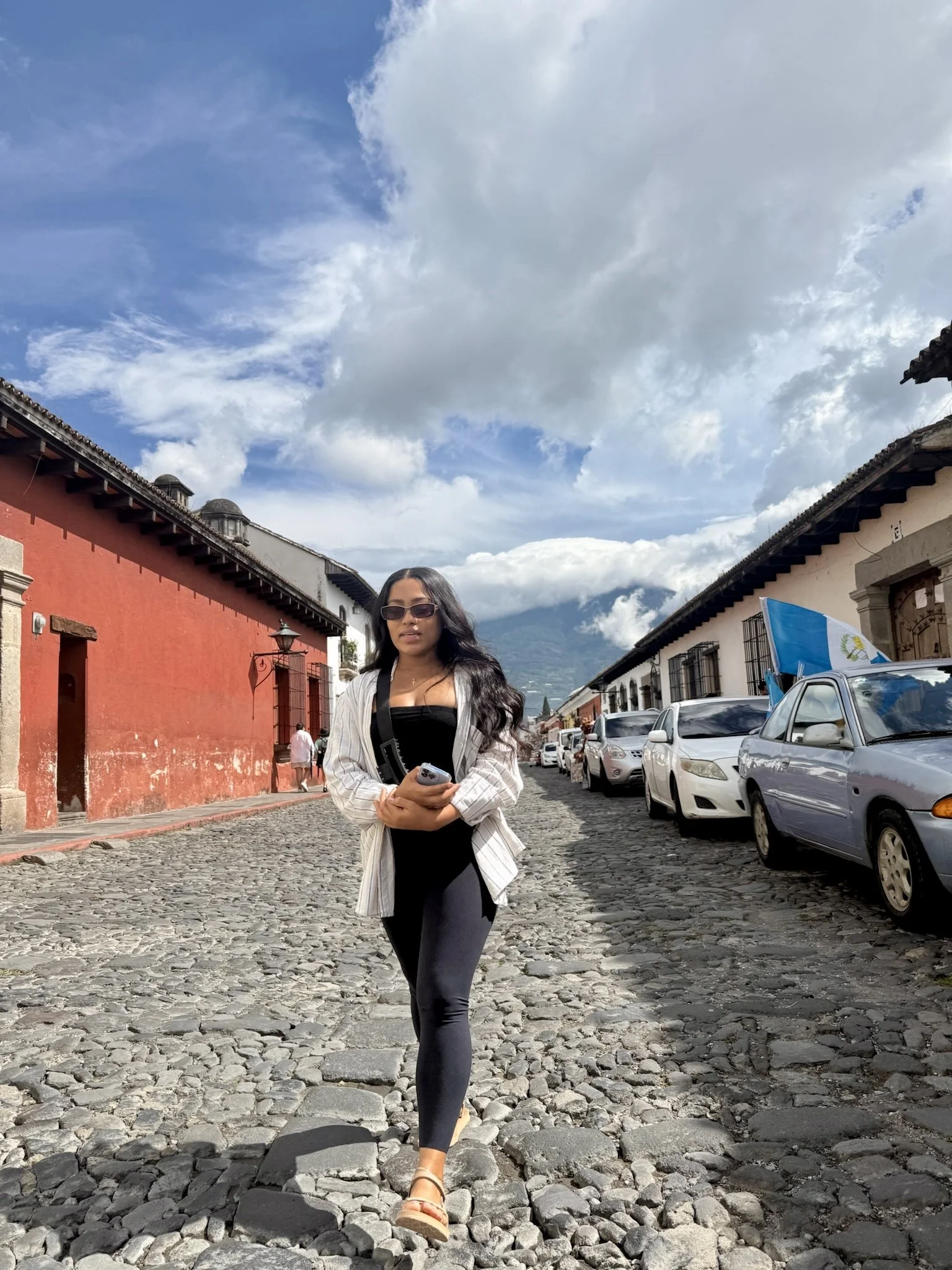 A woman walking on a cobblestone street in a historic town, with colonial-style buildings and parked cars, under a partly cloudy sky, with mountains in the background.