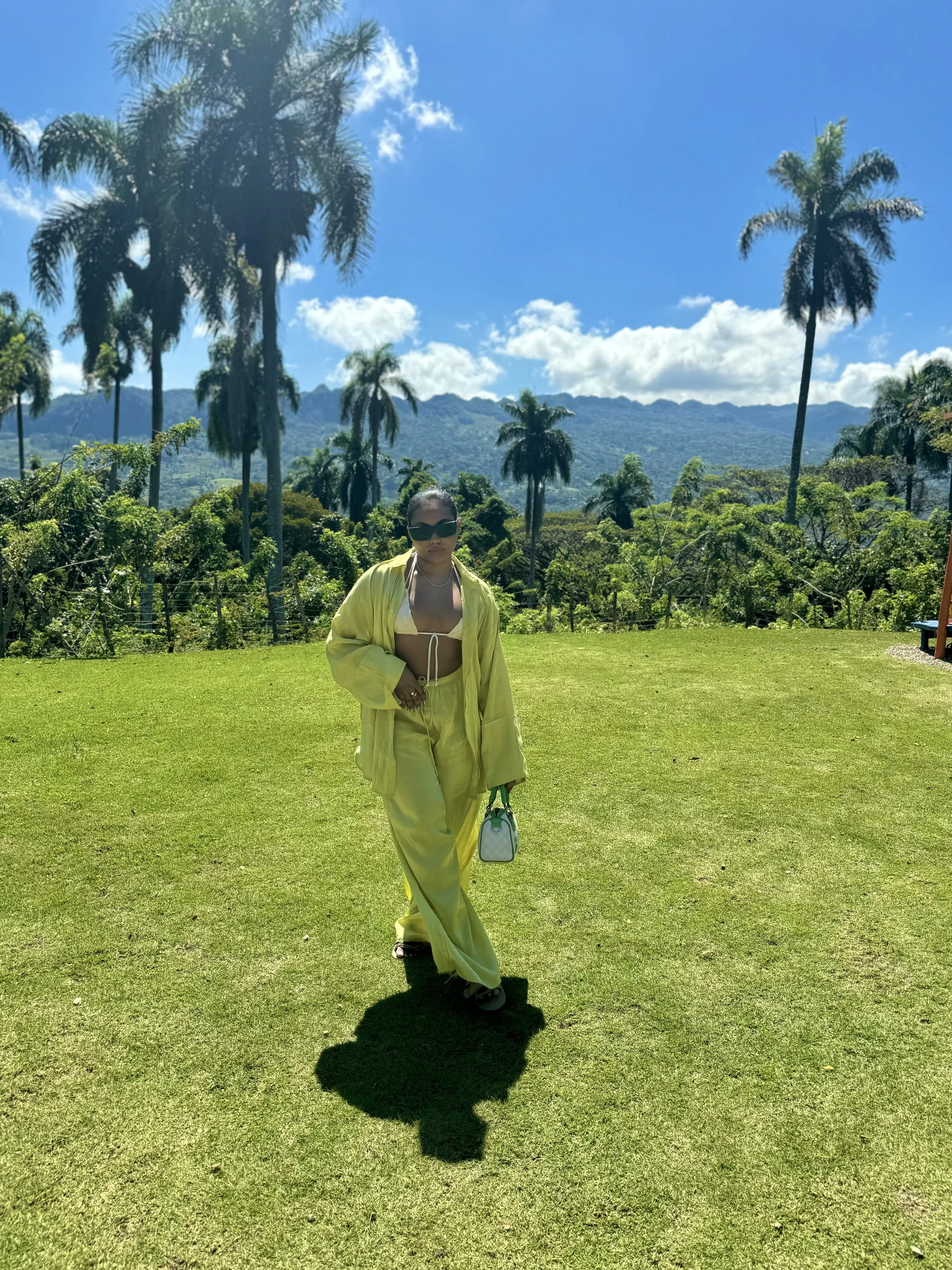 A woman wearing a yellow outfit and sunglasses walking on a grassy field with palm trees and mountain range in the background on a sunny day.