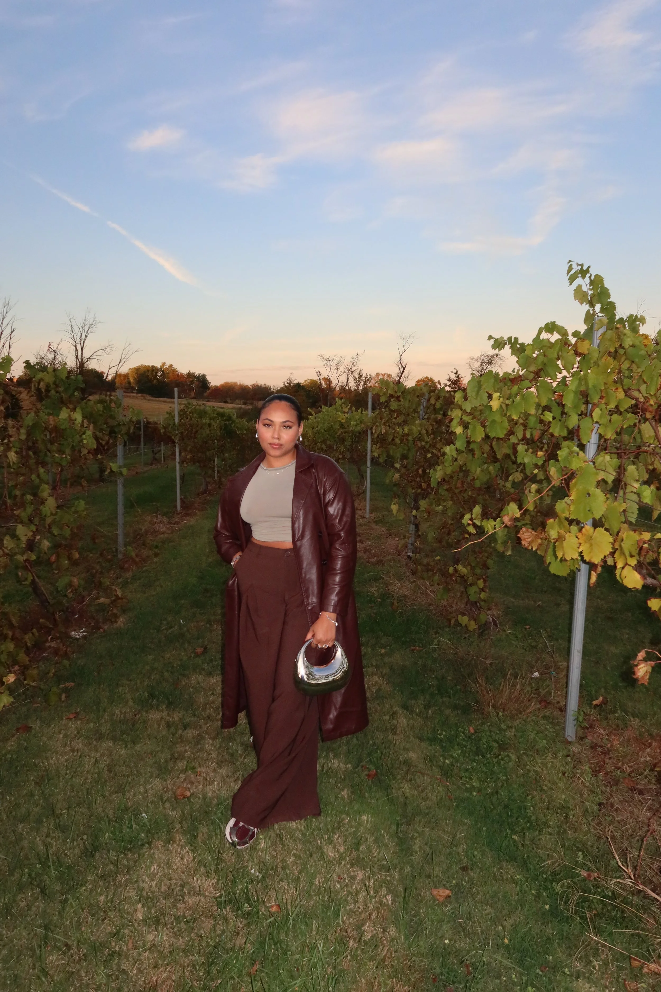 A woman walking through a vineyard at sunset, wearing a long brown leather coat, brown wide-leg pants, a gray top, and holding a shiny metallic handbag.