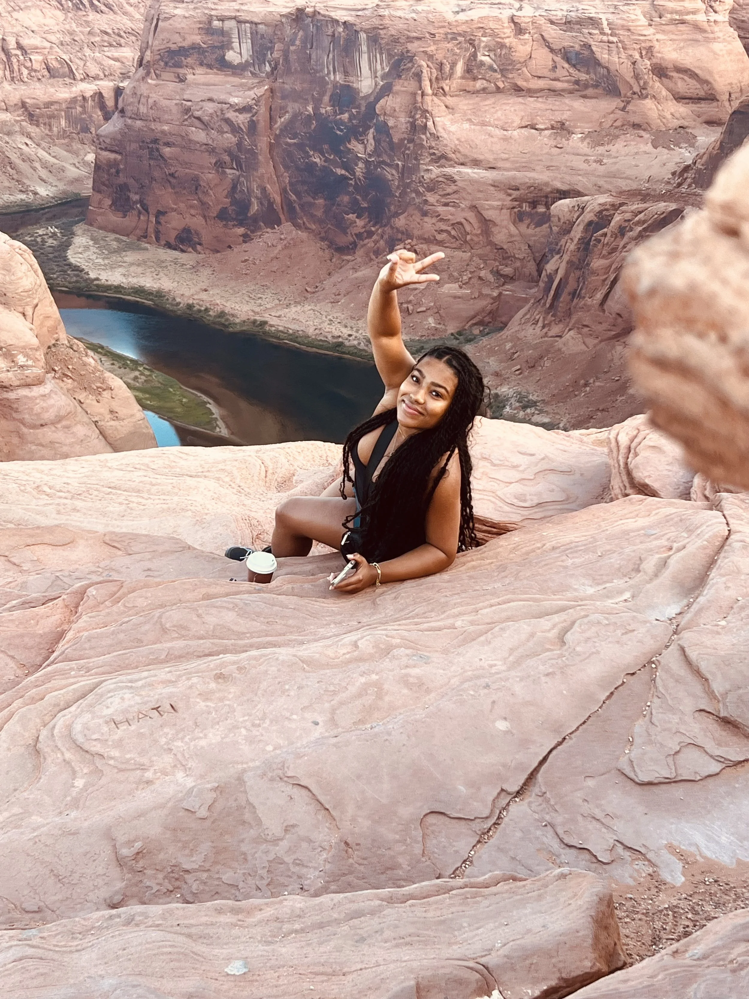 Woman with long dark hair sitting on pink sandstone rock, smiling at camera, with a canyon and river in the background.
