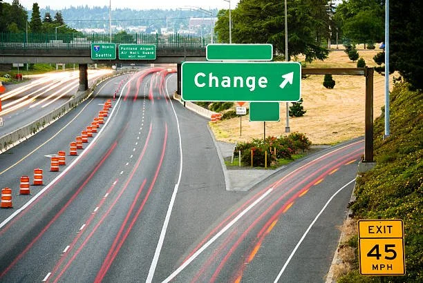 A highway with lanes marked by orange cones and light trails from vehicles, green overhead signs for northbound traffic to Seattle and the airport, and a large green sign indicating a change with an arrow, along with an exit sign for 45 mph.