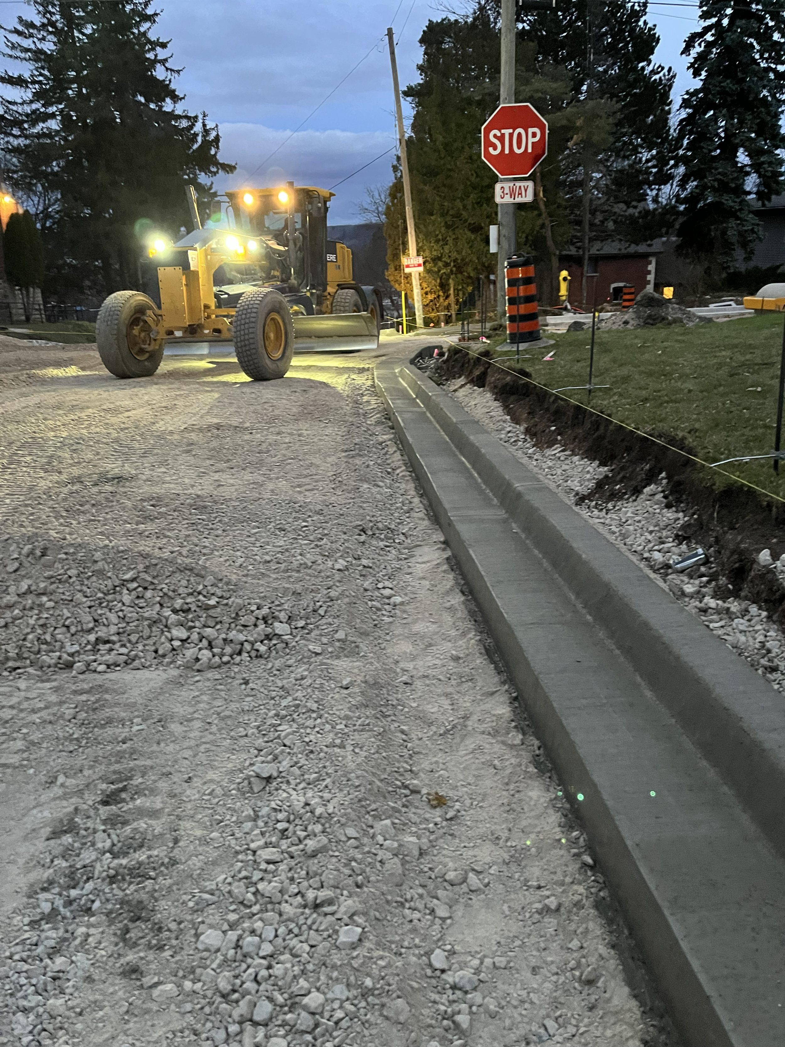 Construction scene at dusk showing a road being paved with a curb installed. A large yellow road roller is parked on the new pavement. Traffic signs, including a stop sign and a 3-way sign, are visible near the construction area. Trees and a building are in the background.