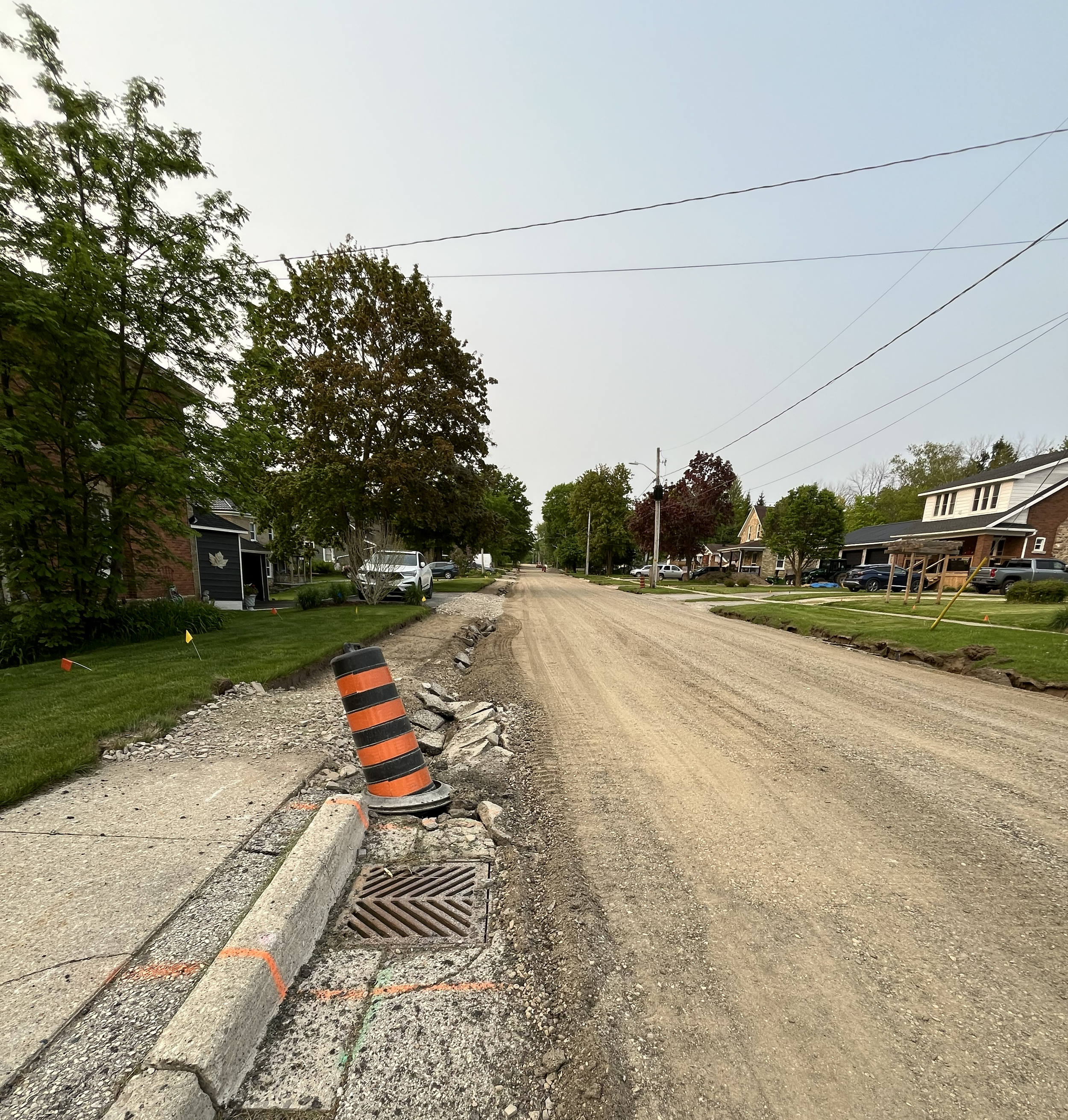 A residential street undergoing road construction with gravel and dirt on the road, orange and black construction barrel, and some construction markers on the grassy areas.