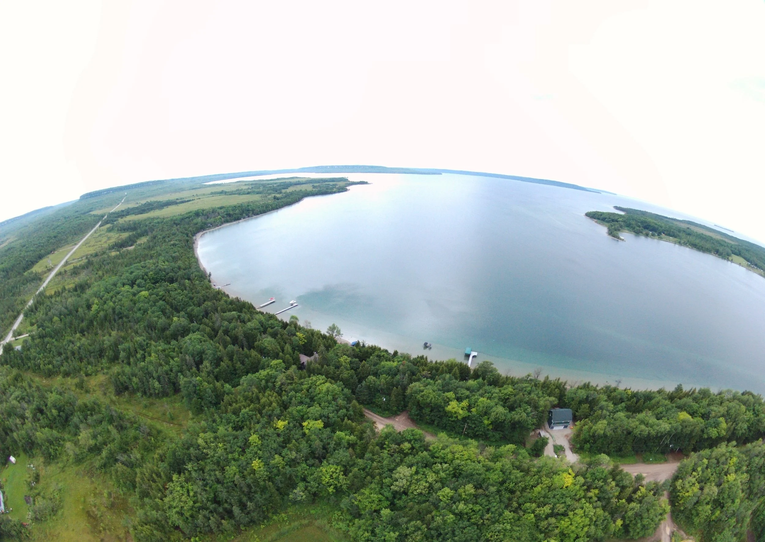 Aerial view of a large lake surrounded by dense green forest and open fields, with a shoreline that has docks and boats, under a cloudy sky.