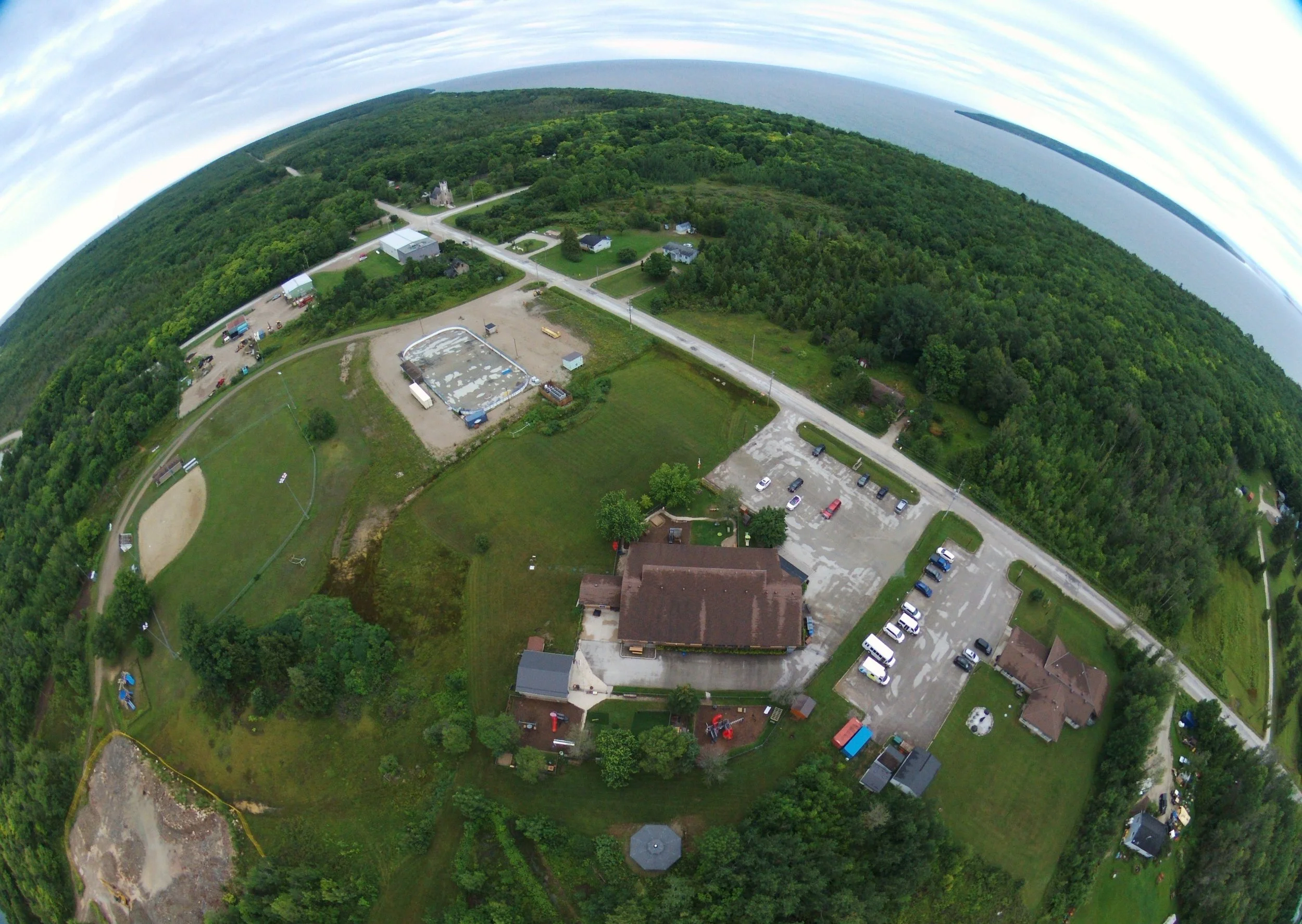 Aerial view of a rural area showing a school, parking lots with cars, a baseball diamond, a swimming pool, wooded areas, and a body of water in the distance.