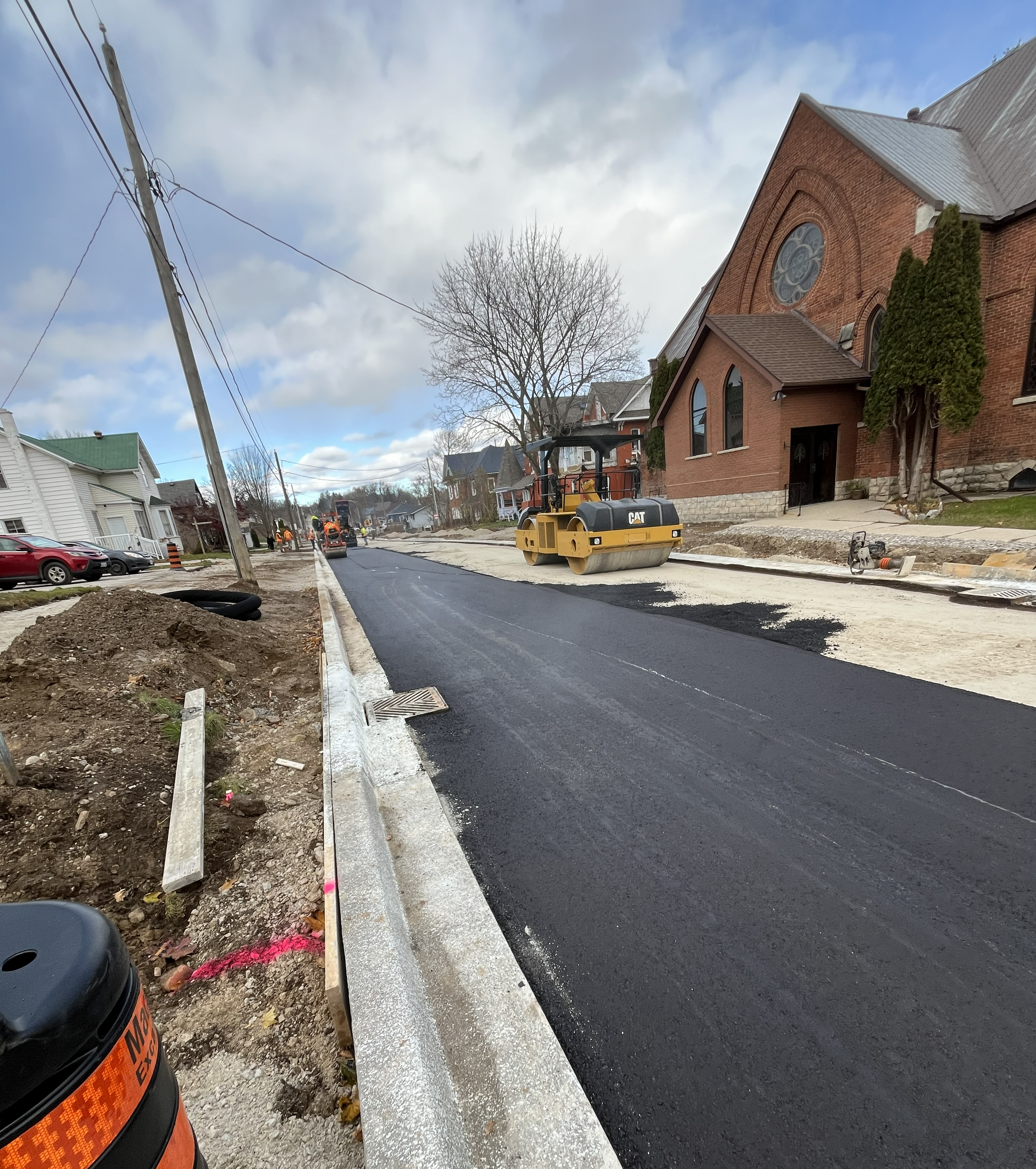 Road construction with a yellow road roller, freshly paved asphalt, and construction workers in the distance on a residential street, with houses and a church in the background.