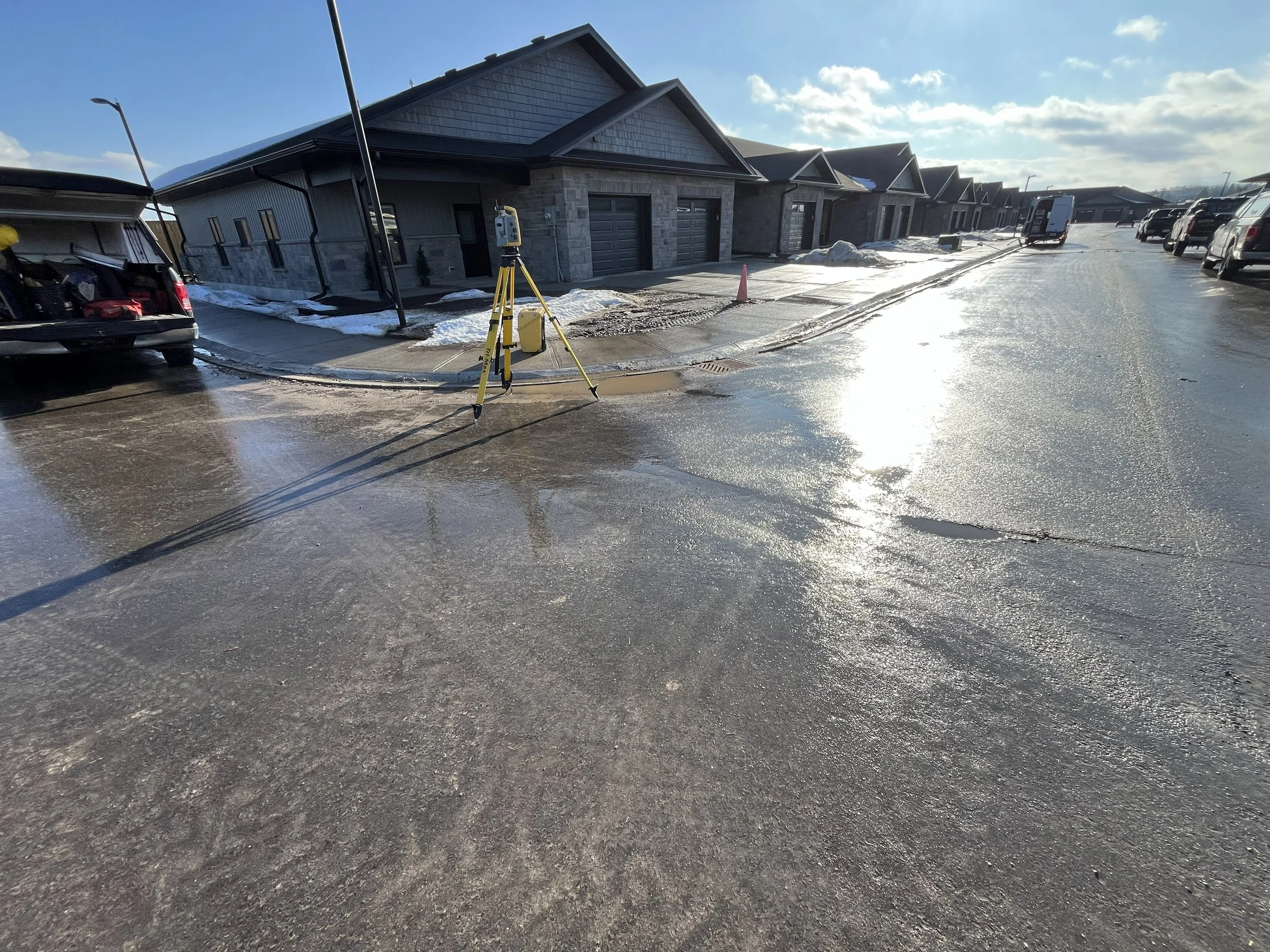 Street scene with newly paved road, parked cars, construction equipment, and houses in the background under a partly cloudy sky.