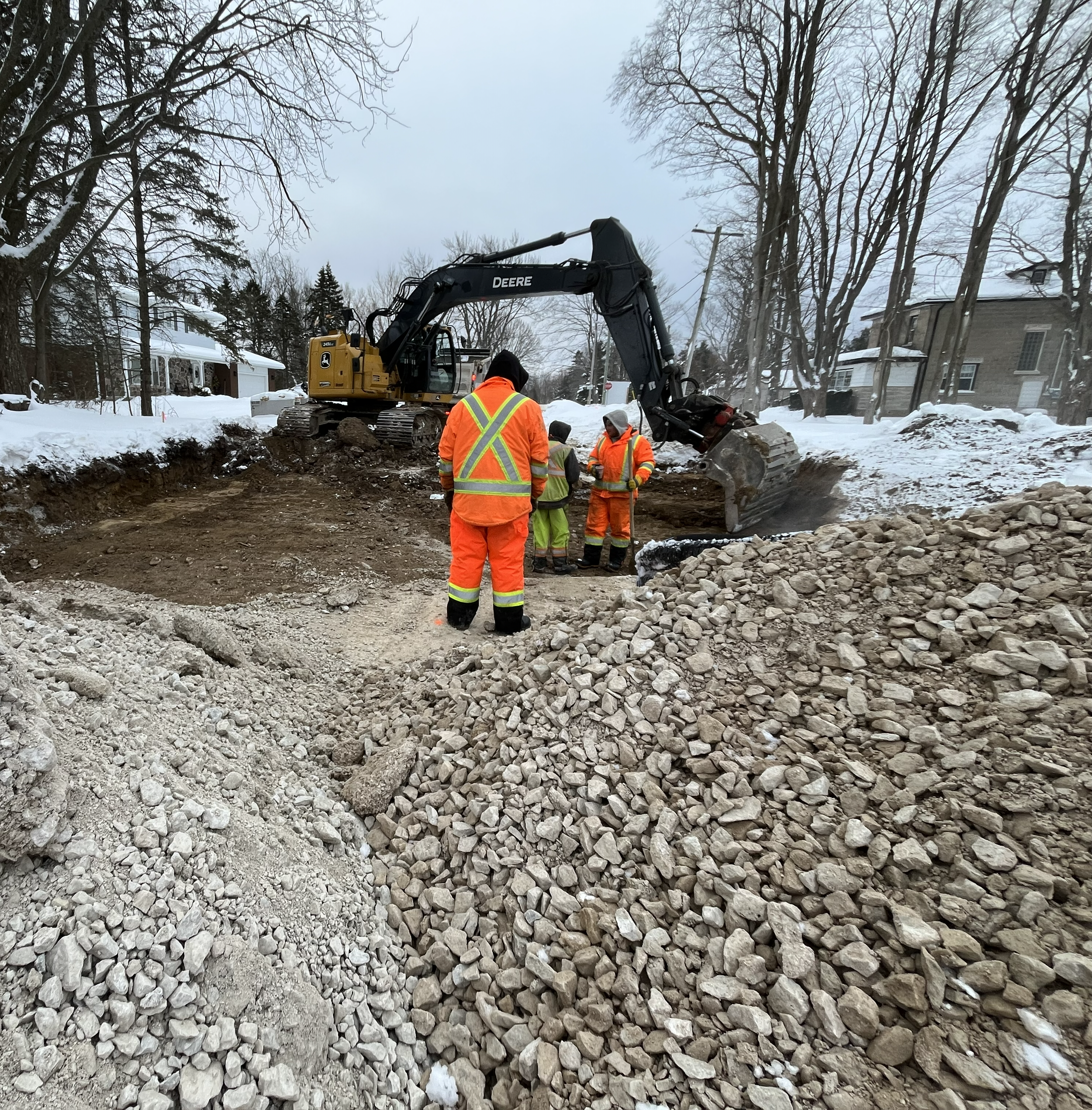 Construction workers in orange safety gear working with an excavator on a snowy site with piles of rocks and dirt, surrounded by leafless trees and residential houses.