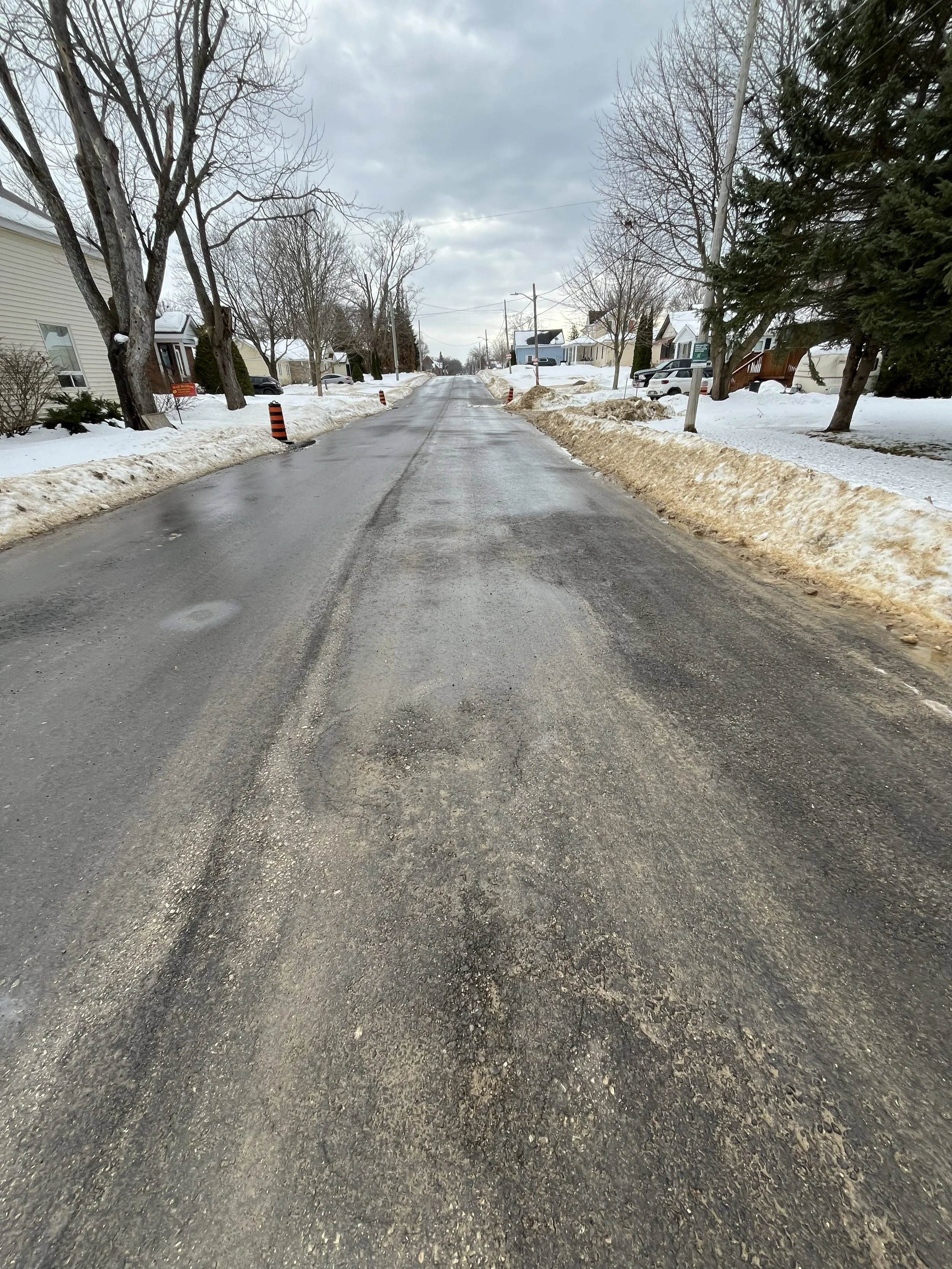 A residential street during winter with snow on the sidewalks and piles of snow along the sides of the road, which is partially paved and has orange and black construction barrels.