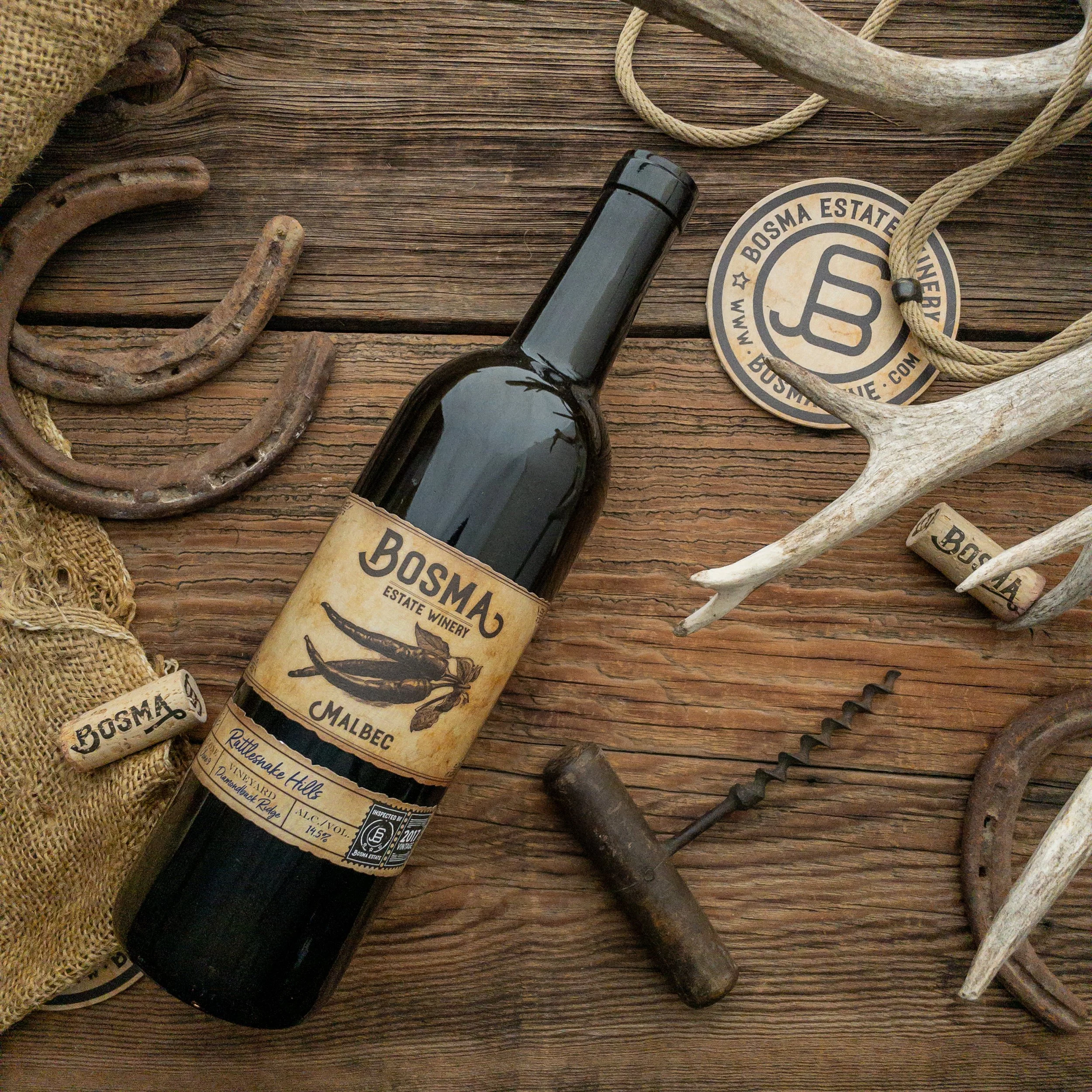 A dark wine bottle with a beige label that has black text reading 'Boss M' and an illustration of a chili pepper, surrounded by rustic objects including an antler, weathered ropes, metal hooks, a corkscrew, and burlap fabric on a wooden surface.