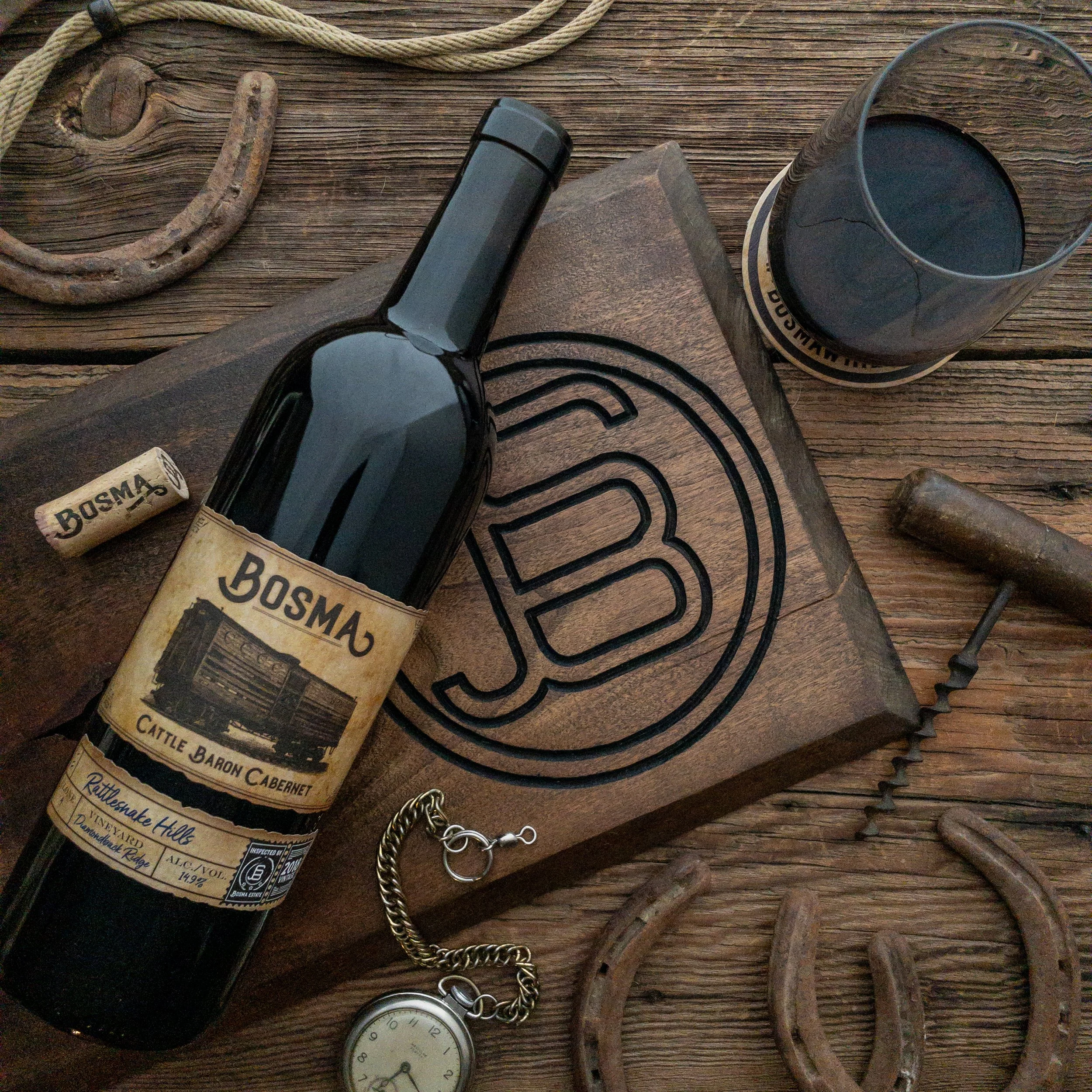 A vintage wooden table holding a black flashlight, two wine corks labeled 'Bosma Cab,' a pocket watch, a horseshoe-shaped nail, a glass of dark liquid, a coiled rope, and a wooden chair.