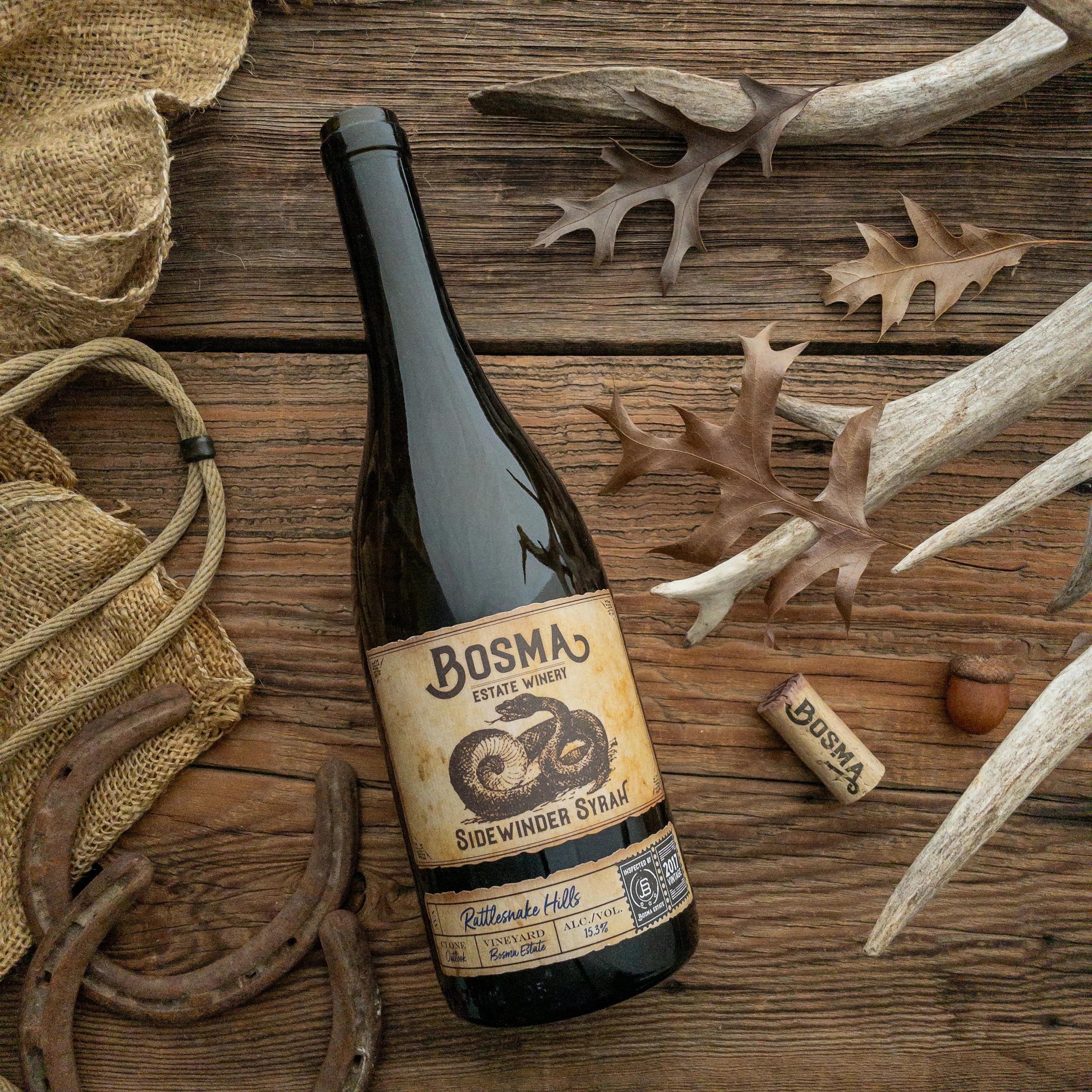 A glass bottle of Bosma Estate Winery wine with a yellow label, a rusty horseshoe, and a piece of burlap on a wooden surface.