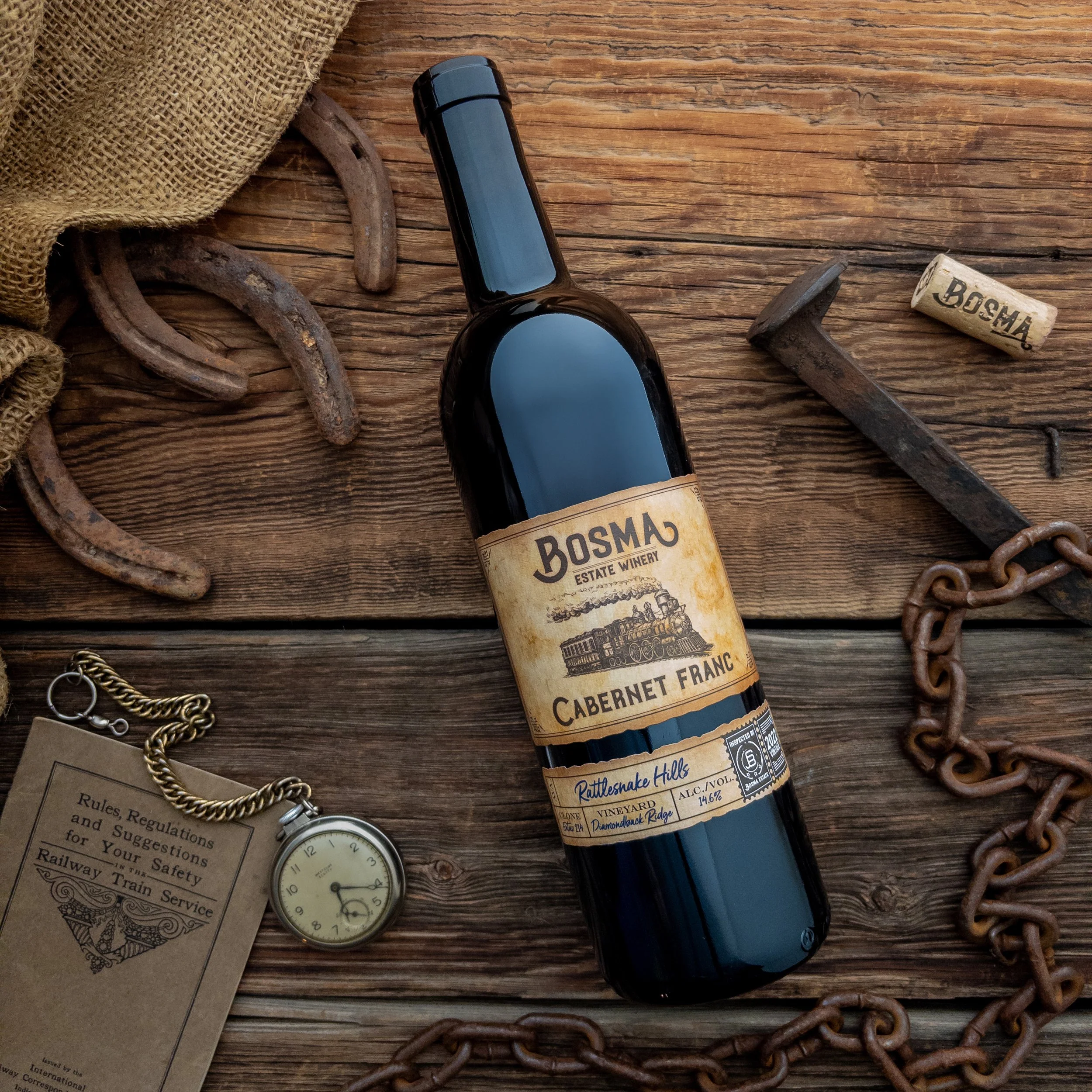 A bottle of Bosmaj estate winery Cabernet Franc wine lies on a rustic wooden surface surrounded by old chains, a vintage pocket watch, a cork, a piece of burlap fabric, and a rule book.