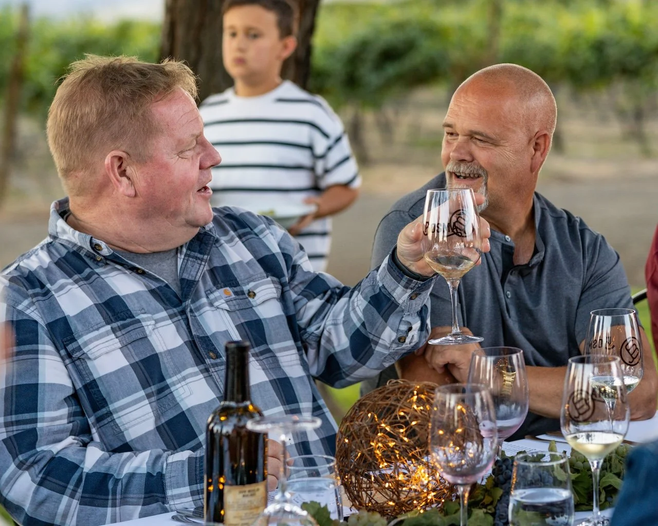Two men enjoying a toast with wine glasses at an outdoor gathering, with a child in the background and a decorative centerpiece on the table.