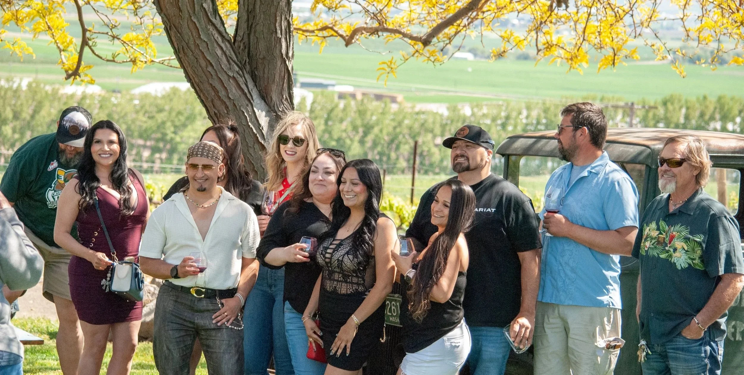Group of people smiling and talking outdoors under a large tree with yellow leaves, holding wine glasses, with a farm or rural landscape in the background.