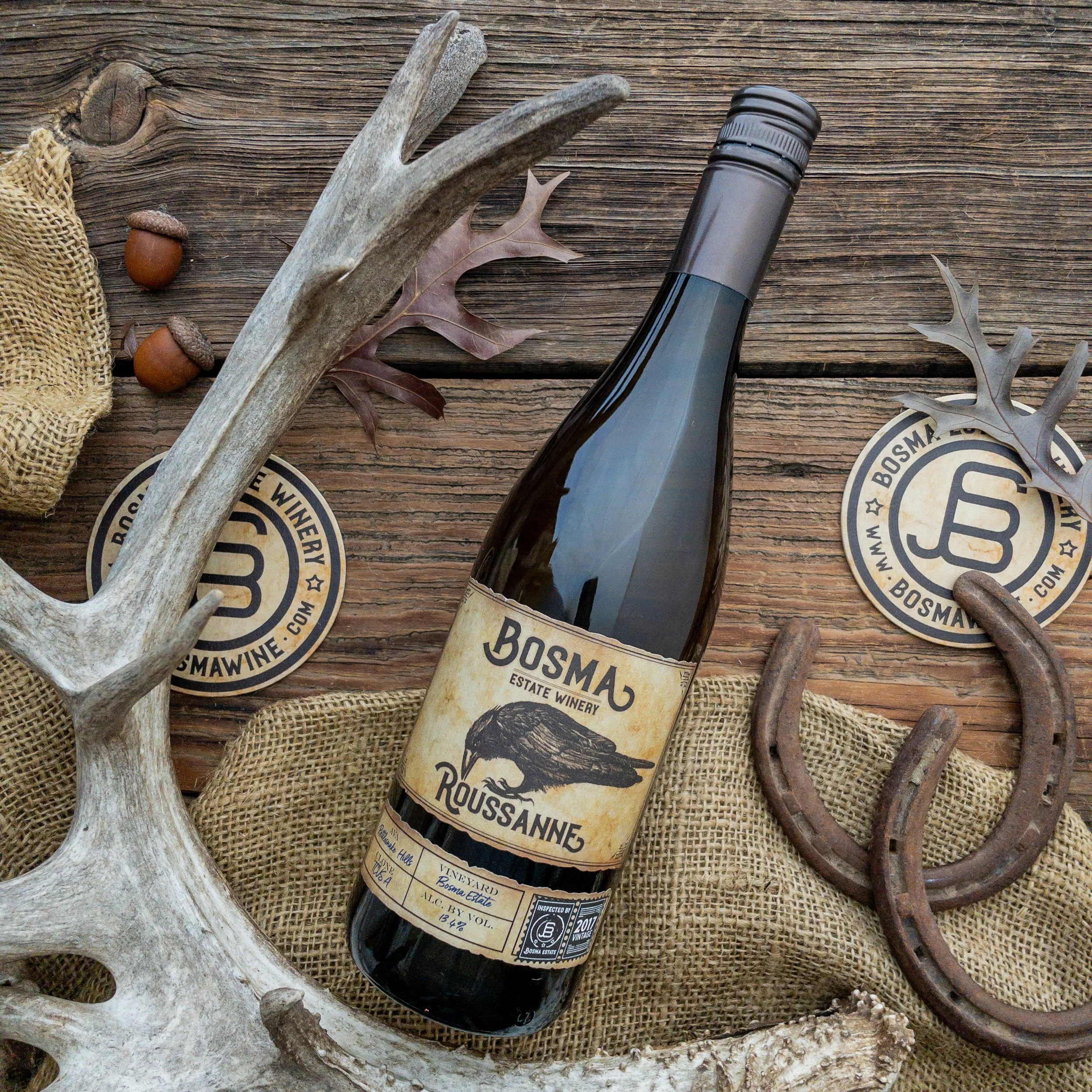 A bottle of Bosma Estate wine on a wooden surface, surrounded by antlers, acorns, and round wooden coasters.
