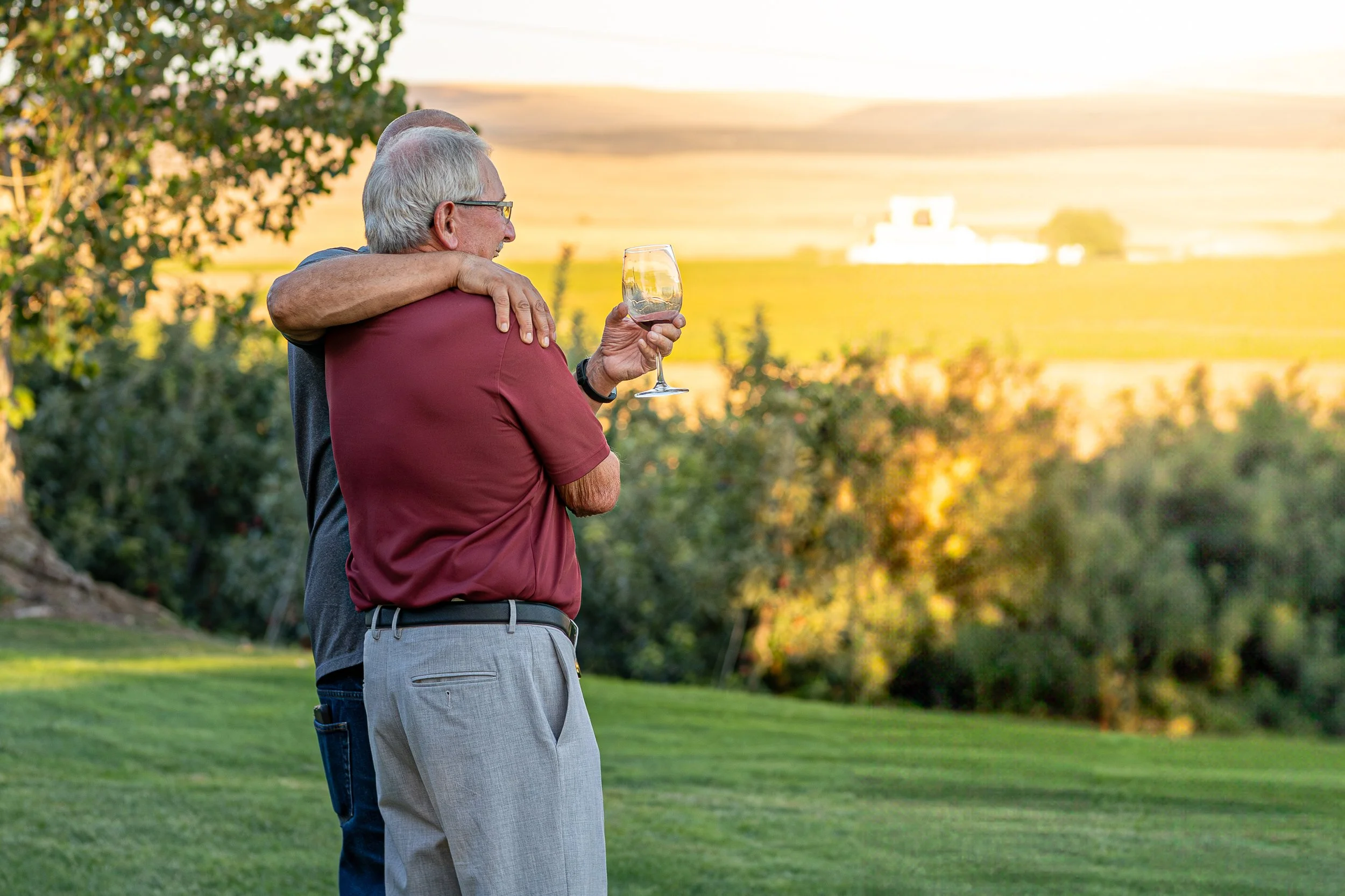 Two elderly men embrace outdoors during sunset, one holding a glass of wine, surrounded by greenery and fields.